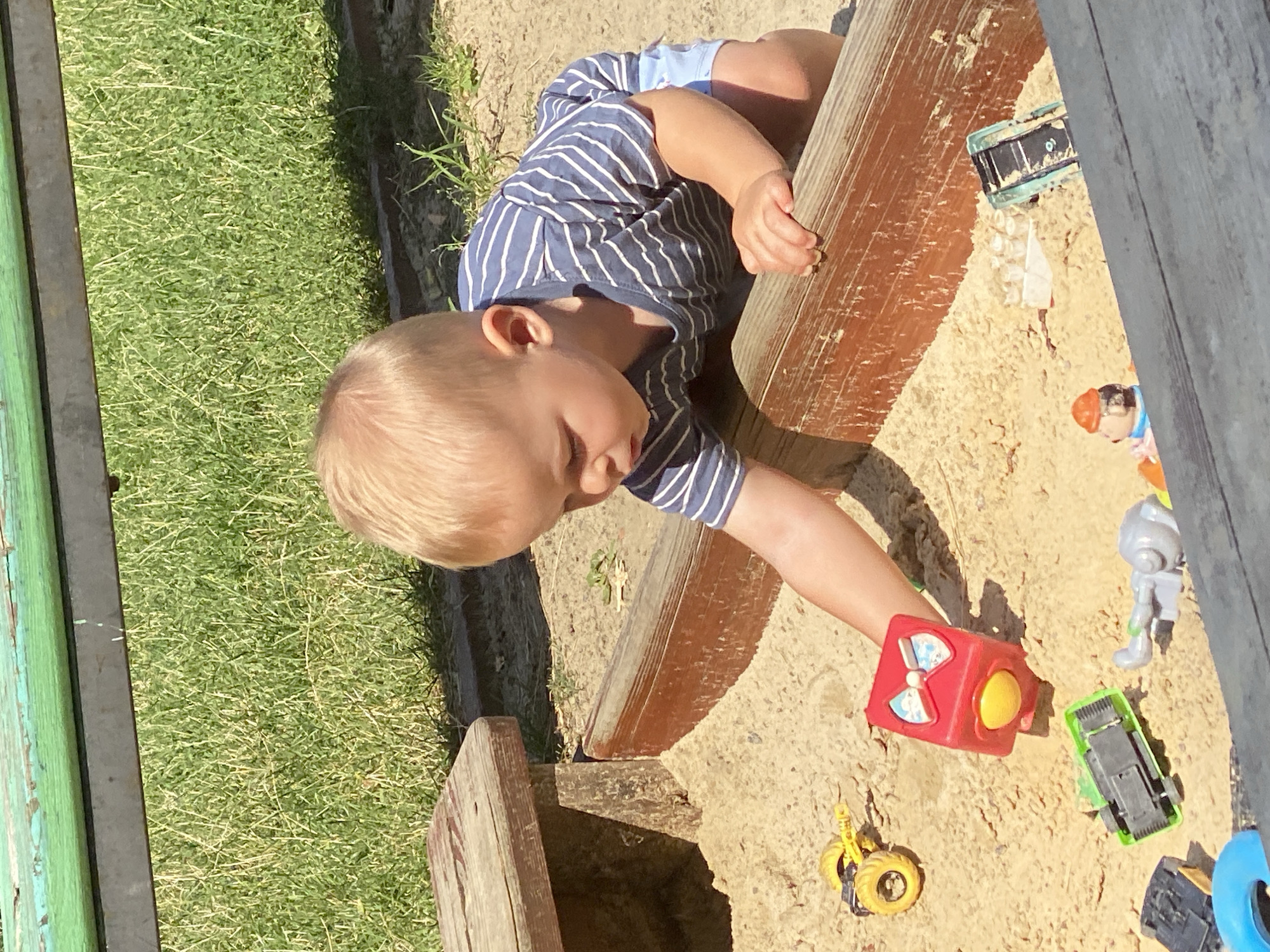 A young boy with blond hair, wearing a blue and white striped t-shirt and light blue shorts, is playing in a sandbox. He is on his knees, reaching for a red toy cube with a yellow button and a bow design on it. Several toy vehicles, including a yellow construction vehicle, a green truck, a black toy car, and a light blue toy truck, are scattered in the sand around him. A small plastic figure wearing an orange hat and blue shirt is also visible in the sandbox. The sandbox has wooden borders. In the background, there is green grass and a wooden bench with a green painted surface. The scene appears to be outdoors on a sunny day, possibly in a park or a playground. The provided image lacks specific details that would allow for identification of the location within the city of Poltava, Ukraine.