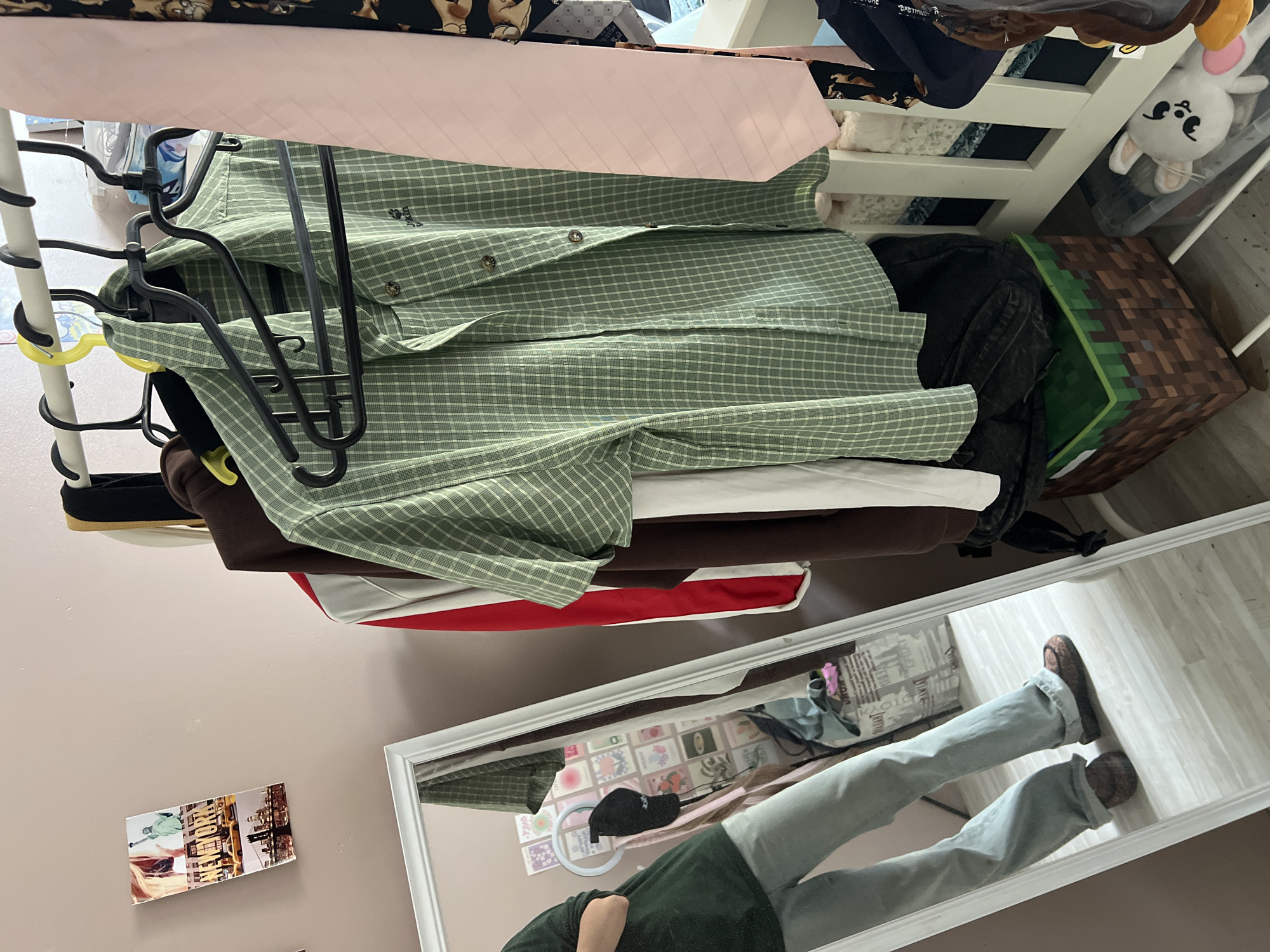 The image depicts an indoor scene, likely a bedroom, featuring a clothing rack with several garments. A person, only their legs and lower torso visible, is reflected in a full-length mirror to the left of the frame. They are wearing light blue, flared jeans and dark, patterned shoes. The mirror also reflects a wall decorated with small square images.
The clothing rack displays a green plaid button-down shirt, a red and white garment, a brown garment, and a light pink tie. To the right of the rack, a white structure, possibly a crib or shelving unit, is partially visible, with more clothing and some stuffed animals. A Minecraft-themed storage box is situated on the floor in front of the clothing rack.
The wall in the background is a pale pink. A postcard featuring New York City imagery is affixed to the wall on the upper left. The floor appears to be light-colored wood. The overall impression is of a personal space, possibly a teenager's room, with a focus on clothing and personal items. The location is specified as Мухавецкі сельскі Савет, Belarus, though there are no visual cues within the image to confirm this specific location.