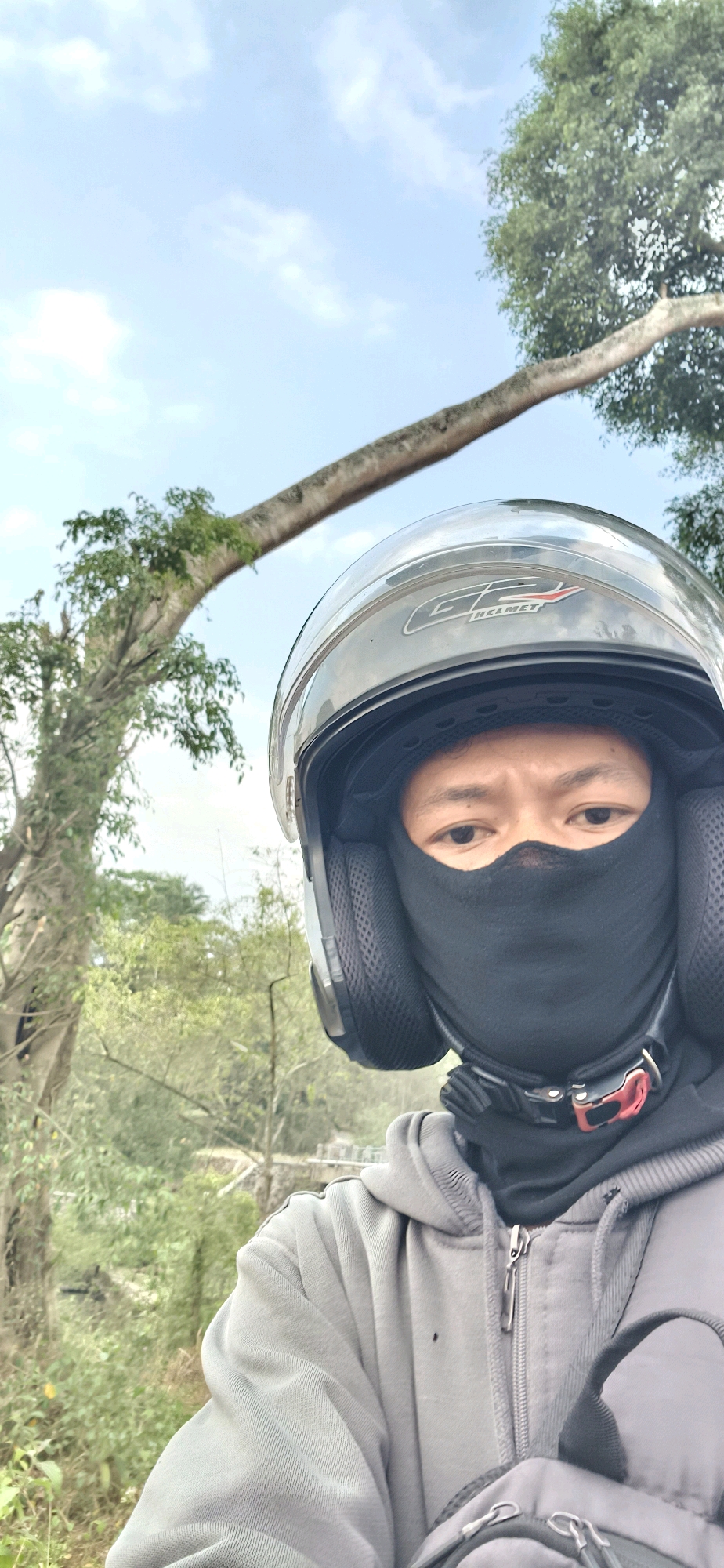 In Tengahan, Indonesia, a person wearing a motorcycle helmet and a black face covering is captured in a selfie. The person is wearing a gray hooded jacket. The background is filled with lush green trees and a light blue sky with some clouds. A large tree branch extends diagonally across the upper portion of the image. The overall scene suggests an outdoor setting, possibly a rural or natural area within Tengahan.