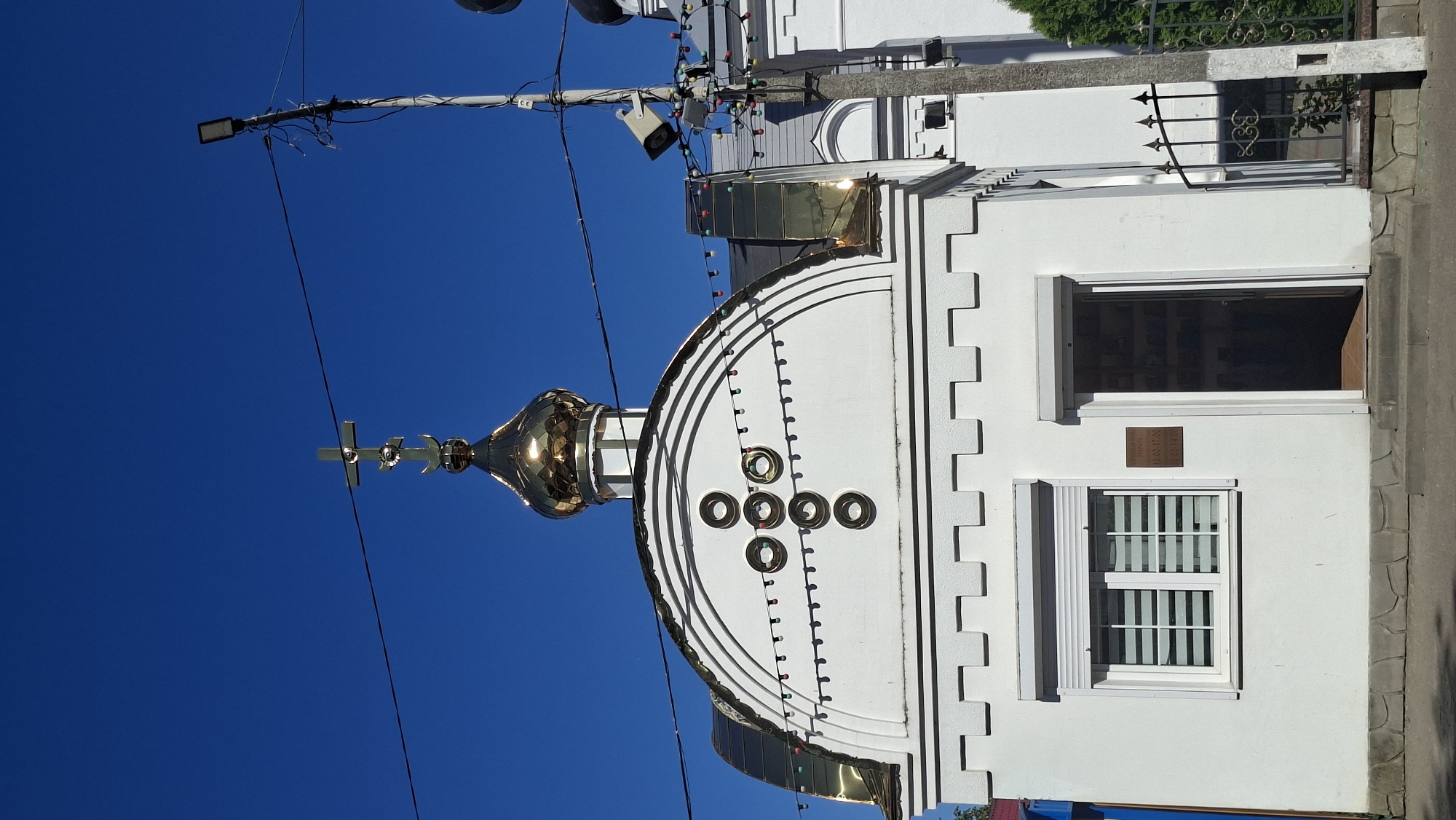The image depicts the facade of a white church building with a golden dome topped by a cross. The church has a semicircular arched facade decorated with a large cross formed by four circular ornaments. A string of multi-colored lights is draped across the facade, just below the decorative cross. To the left of the entrance, there is a window with blinds. The entrance itself is an open doorway. Above the doorway, a small sign is mounted. The building appears to be situated in an urban environment, with a clear blue sky overhead. A utility pole with electrical wires and a security camera is visible to the right of the church. A metal fence is also present. The overall scene is a daytime exterior shot, likely in a well-lit area. While the image is identified as being in Pastavy, Belarus, no specific landmarks or contextual clues within the image itself pinpoint this exact location.