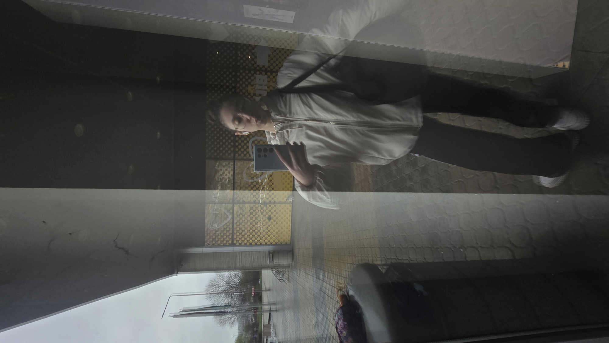 A reflection of a person taking a picture of themselves is visible in a glass window. The person is wearing a white jacket, dark pants, and white shoes. They are holding a smartphone. The reflection also shows a street scene, including a sidewalk with a patterned surface, bare trees, and what appears to be part of an outdoor seating area. The location is Stara Zagora, Bulgaria.