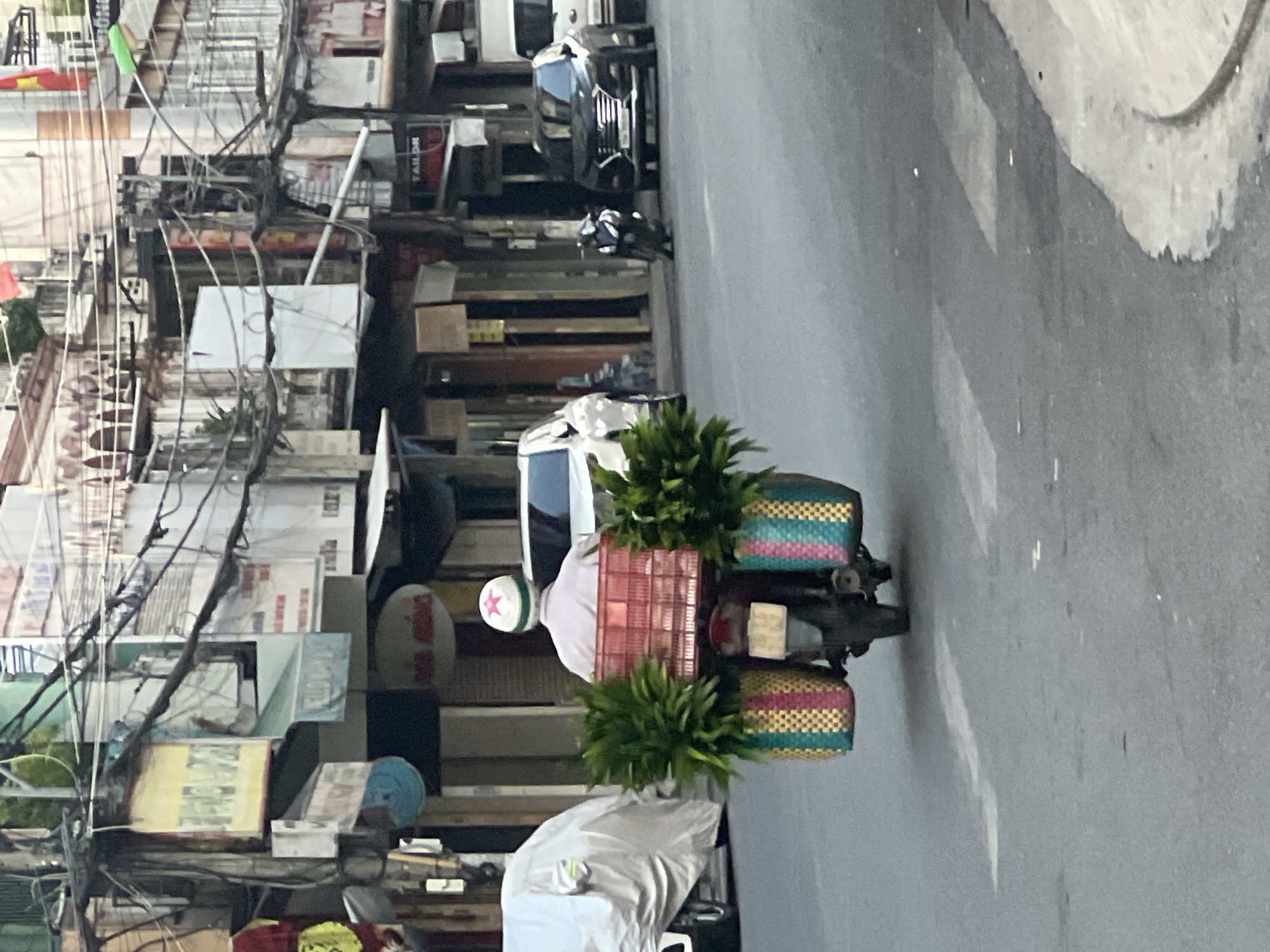 In Nha Trang, Vietnam, a person wearing a white helmet with a red star rides a motorcycle. They are carrying a large red crate filled with plants on the back of the motorcycle, with two large woven baskets on either side of the crate also filled with plants. The scene is set on a street with buildings and power lines lining the background. A white car is parked to the left of the motorcycle, and a black SUV and a white van are visible further down the street. The motorcycle appears to be moving forward on the asphalt road.