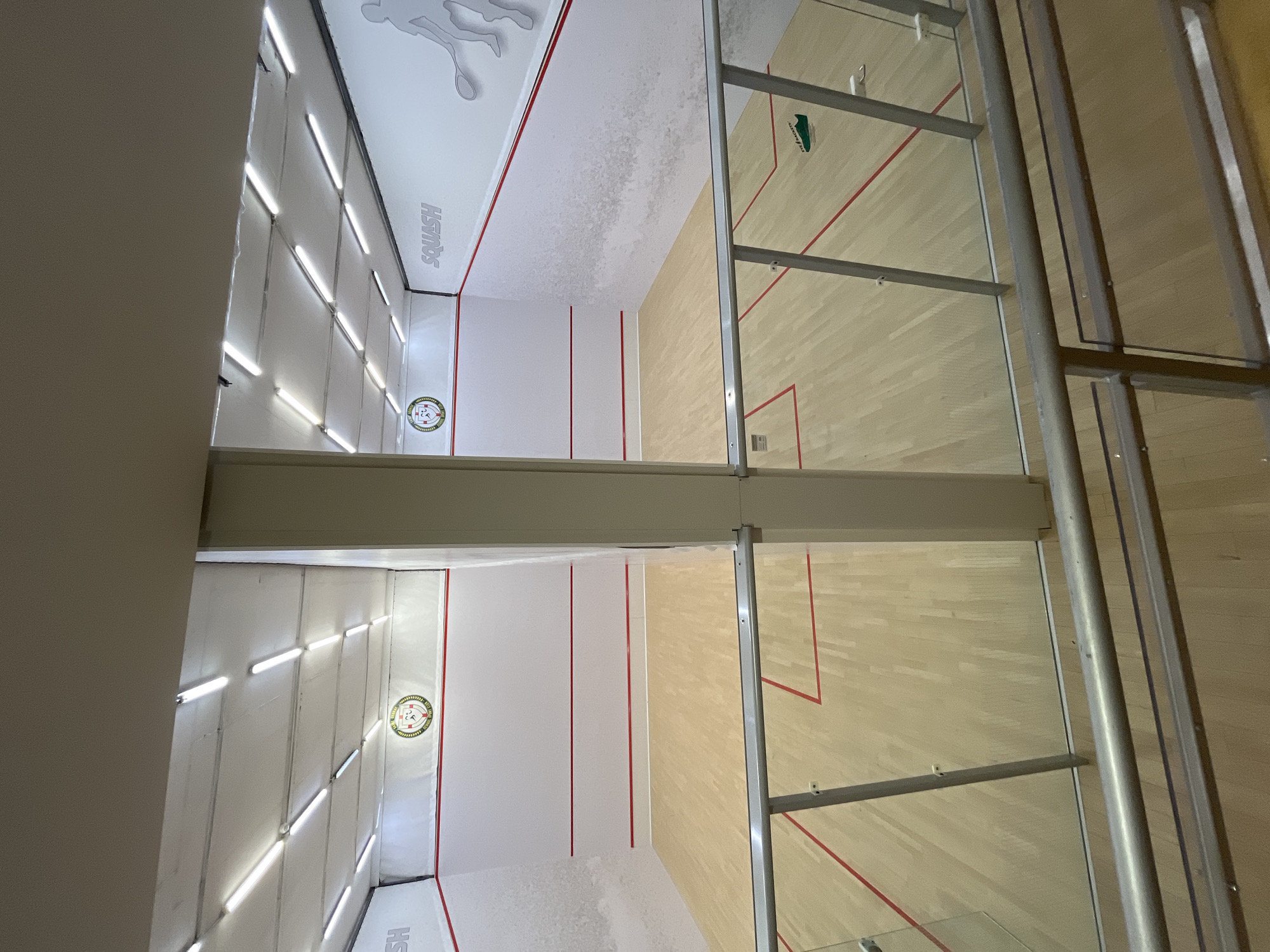 The image shows an interior view of two adjacent squash courts separated by a central pillar. Both courts have polished wooden floors marked with red lines indicating the playing boundaries. The walls are white with red lines running horizontally across them, and a red diagonal line from the front wall to the side wall. A round emblem is visible on the left wall of the left court, and the word "SQUASH" is stenciled on the right wall of the right court, along with a silhouette of a squash player. The courts are illuminated by rows of fluorescent lights on the ceiling. A glass partition with metal railings runs along the foreground, suggesting this view is from a spectator area or elevated walkway. A green shoe with the text "EYE RACKETS" is visible on the floor of the right court. The overall environment appears to be a sports facility.