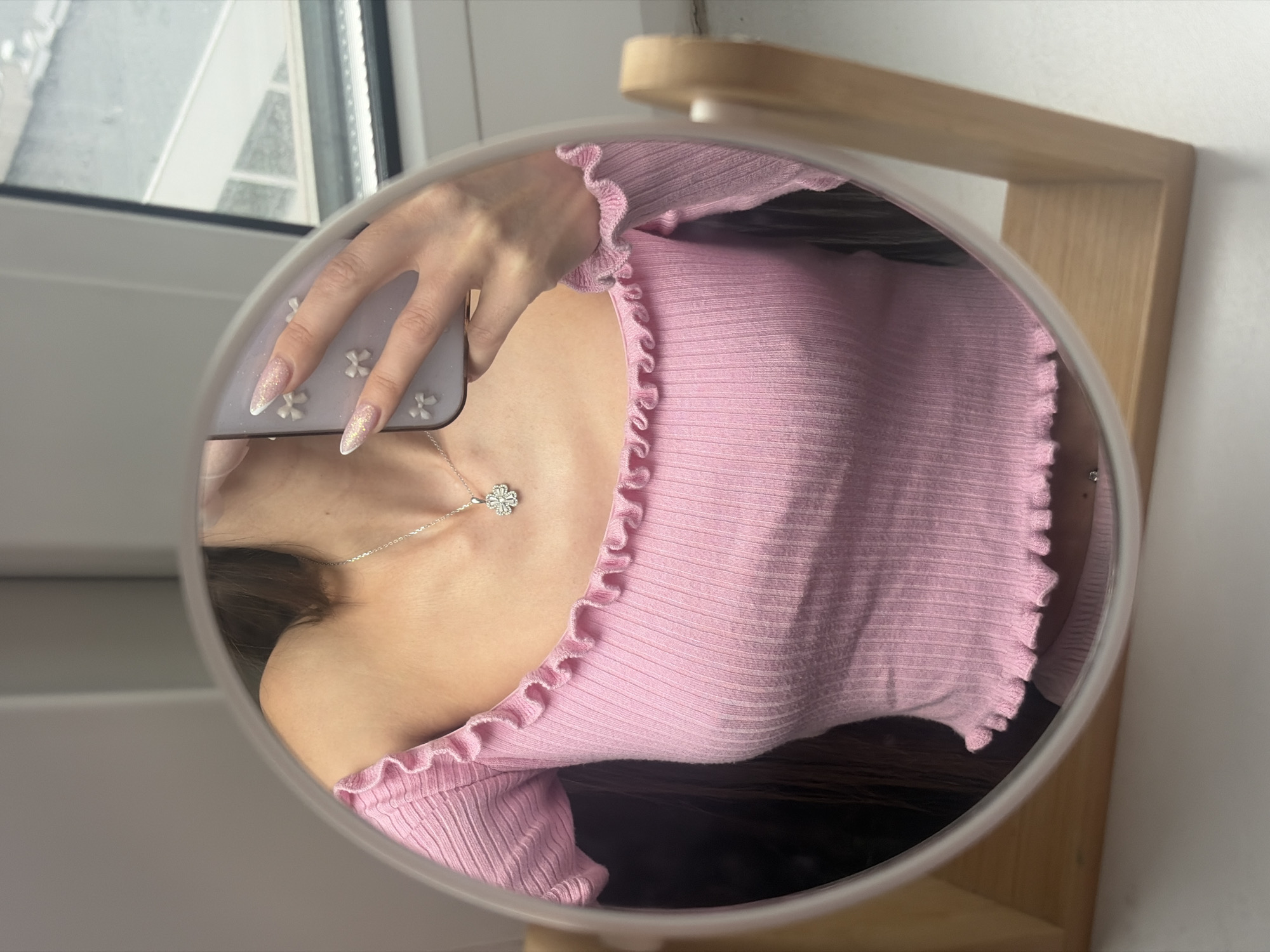 A mirror reflects a person wearing a pink, ruffled-neck shirt and a silver clover necklace. The person holds a mobile phone in their hand, and its case is decorated with floral designs. The background includes a window. It is not possible to determine the location as St. Petersburg, Russia, from the visual information alone.