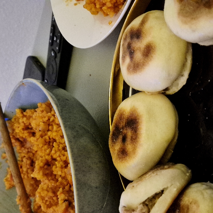 A grey bowl filled with vibrant orange rice and a wooden spoon is partially visible. A small white plate holding similar rice is nearby. A gold tray holds a stack of slightly toasted English muffins. One muffin appears halved, revealing its interior. A black remote control rests in the background.