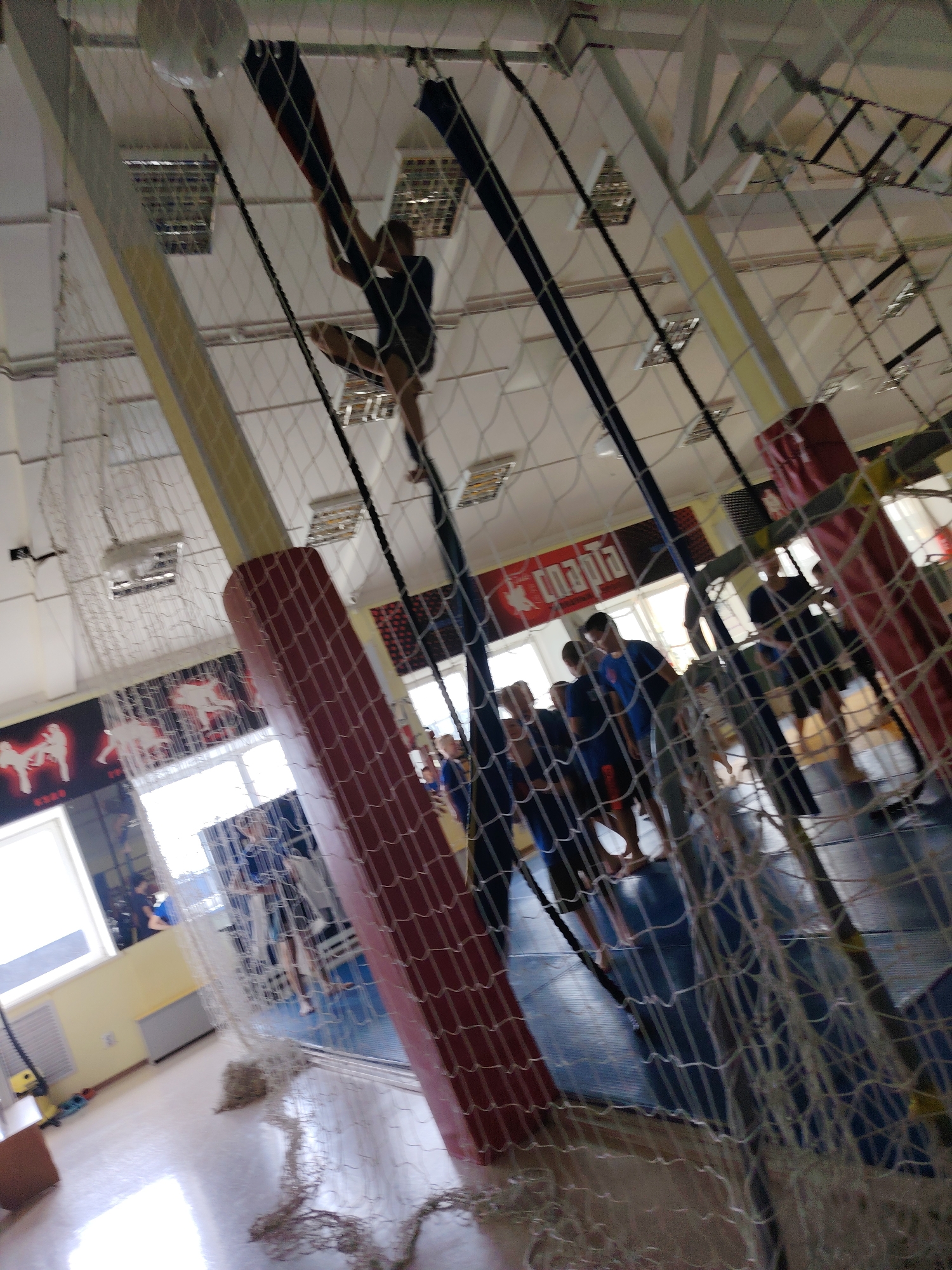 The image captures an indoor sports facility in Chaikovsky, Russia. In the foreground, a net hangs in front of the camera, obscuring some of the view. Behind the net, a person is seen climbing on what appears to be a rope or climbing apparatus suspended from the ceiling. The person is wearing dark clothing. Further into the facility, several other individuals are visible, some standing and others in motion, on a blue mat. The environment includes a white ceiling with lighting fixtures and structural beams, red padding on a vertical support, and windows allowing natural light. Two posters are visible on the wall, one with silhouetted figures engaged in martial arts and another with Cyrillic text. The overall impression is that of a gymnasium or training center.