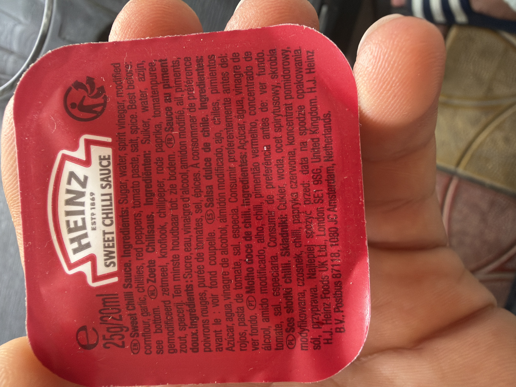 A close-up shot captures a person's hand holding a small, red Heinz Sweet Chilli Sauce sachet. The hand's thumb and fingers are prominently featured, displaying visible skin texture and fingerprints. The sachet is slightly bent as it's held.
The packaging is red with white and black text. The "HEINZ" logo with "ESTP 1869" is visible, along with "SWEET CHILLI SAUCE" and "25g/20ml." A recycling symbol (a person dropping an item into a bin with arrows) is printed near the top left of the sachet.
Extensive product information is printed on the sachet in multiple languages, including English, Dutch, French, Spanish, Portuguese, and Polish. Key visible text includes "Sweet Chilli Sauce. Ingredients: Sugar, water, spirit vinegar, modified cornflour, garlic, chillies, red peppers, tomato paste, salt, spice. Best before: see bottom." Similar ingredient lists are provided in other languages. Manufacturer details are also present: "H.J. Heinz Foods UK Ltd., London SE1 9SG, United Kingdom. H.J. Heinz B.V., Postbus 87118, 1080 JC Amsterdam, Netherlands."
The background is blurred, showing indistinct patterns that suggest an indoor setting, possibly fabric or upholstery. There are no discernible time of day, weather, or emotional cues. The image focuses almost exclusively on the hand and the product packaging.