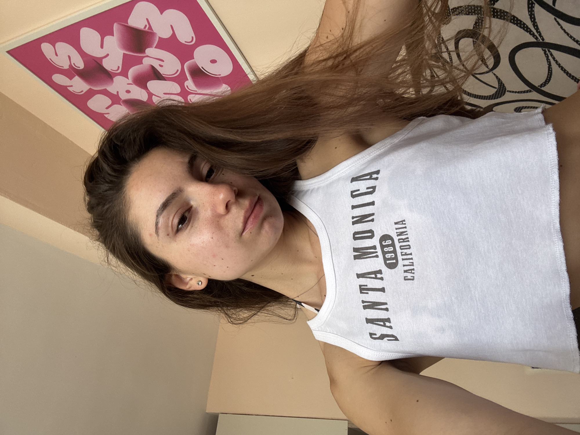 The media file is a portrait of a young woman taken indoors. She has long, brown hair and appears to be in her late teens or early twenties. She is wearing a white tank top with black text that reads "SANTA MONICA 1986 CALIFORNIA". The background includes a pink poster with bubble-like white text and a patterned wallpaper. The lighting suggests it is daytime, and there are no indications of specific weather conditions. The woman's expression is neutral, and she is looking towards the camera. She has a small silver stud earring in her visible ear. The overall scene is a casual, personal snapshot.