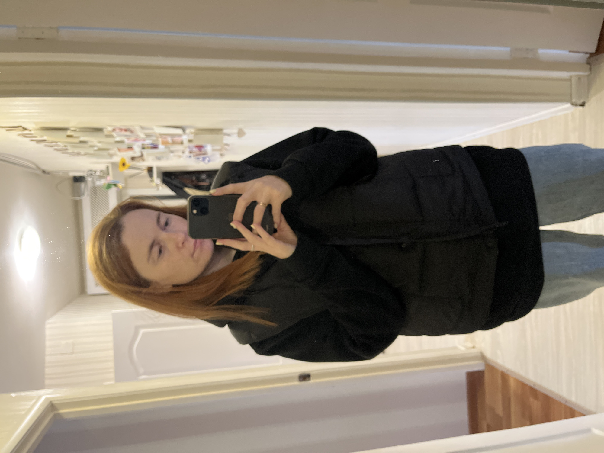A selfie of a light-skinned person with red hair wearing a black hoodie and puffer vest, along with blue jeans, is reflected in a mirror. The background indicates a hallway, and a collage of papers is visible on the wall. The location is Chaikovsky, Russia.