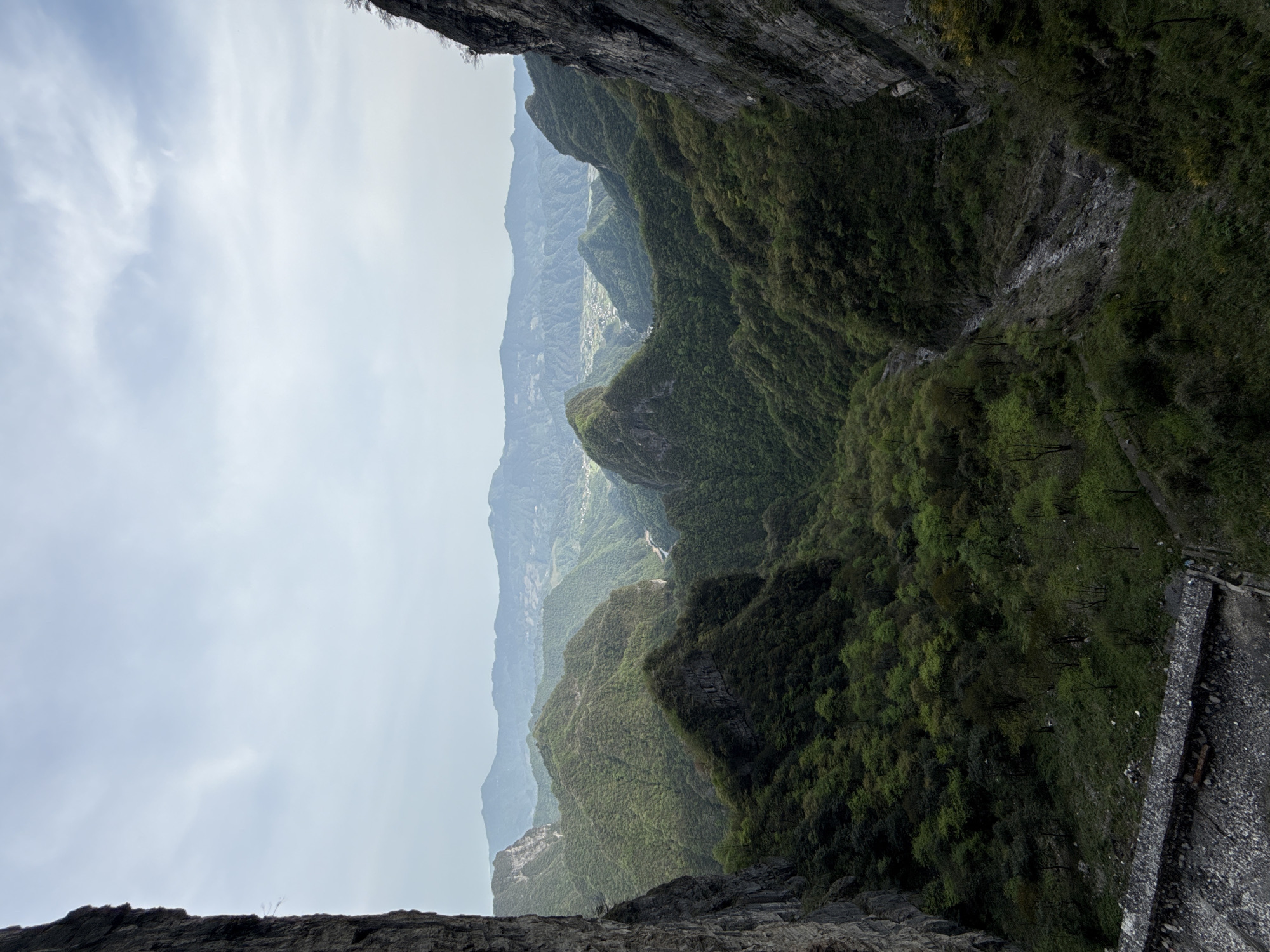 The image shows a steep, mountainous landscape in Yongding District, China. The viewpoint is from a high cliff, looking down into a deep valley. The sky is overcast. The valley is filled with layered, green mountains covered in dense vegetation. A narrow, concrete path or wall is visible along the edge of one of the mountains. The foreground shows the rocky cliffs from which the image is taken.