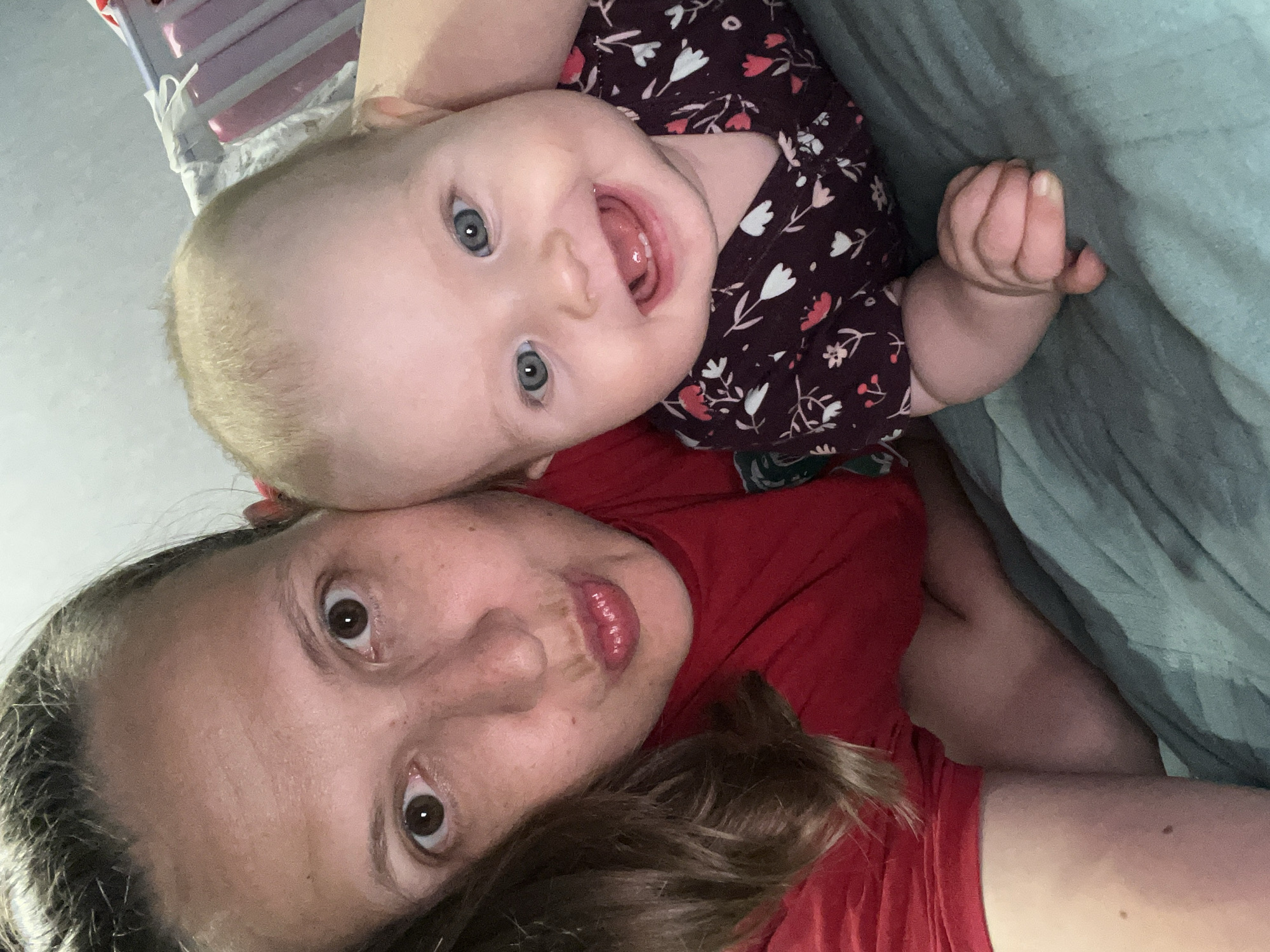The image shows a selfie of a woman and a baby. The woman is lying down, wearing a red shirt, and has brown hair. The baby is positioned above the woman, wearing a dark-colored floral print onesie, and is looking at the camera with an open mouth. A teal-colored sheet or blanket is visible in the background. A partial view of a baby crib is visible in the top left of the image. The location is not identifiable from the image content.