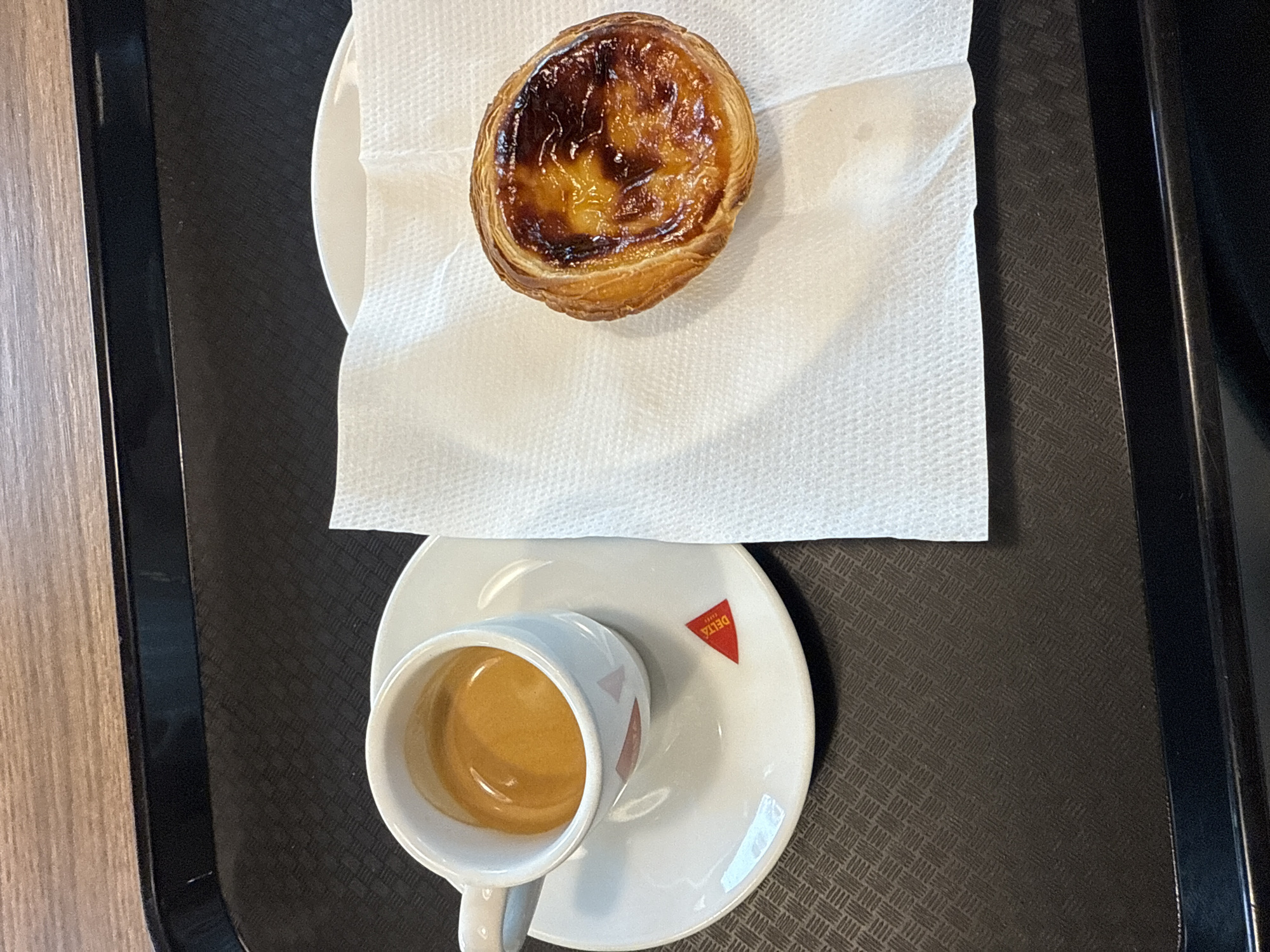 On a black tray, an espresso in a white "Delta" cup and saucer is positioned below a Portuguese custard tart (Pastel de nata) served on a small plate with a white paper napkin. The items appear to be situated on a table with light-colored wood grain visible on the left edge of the image. The location is Pombal, Portugal.