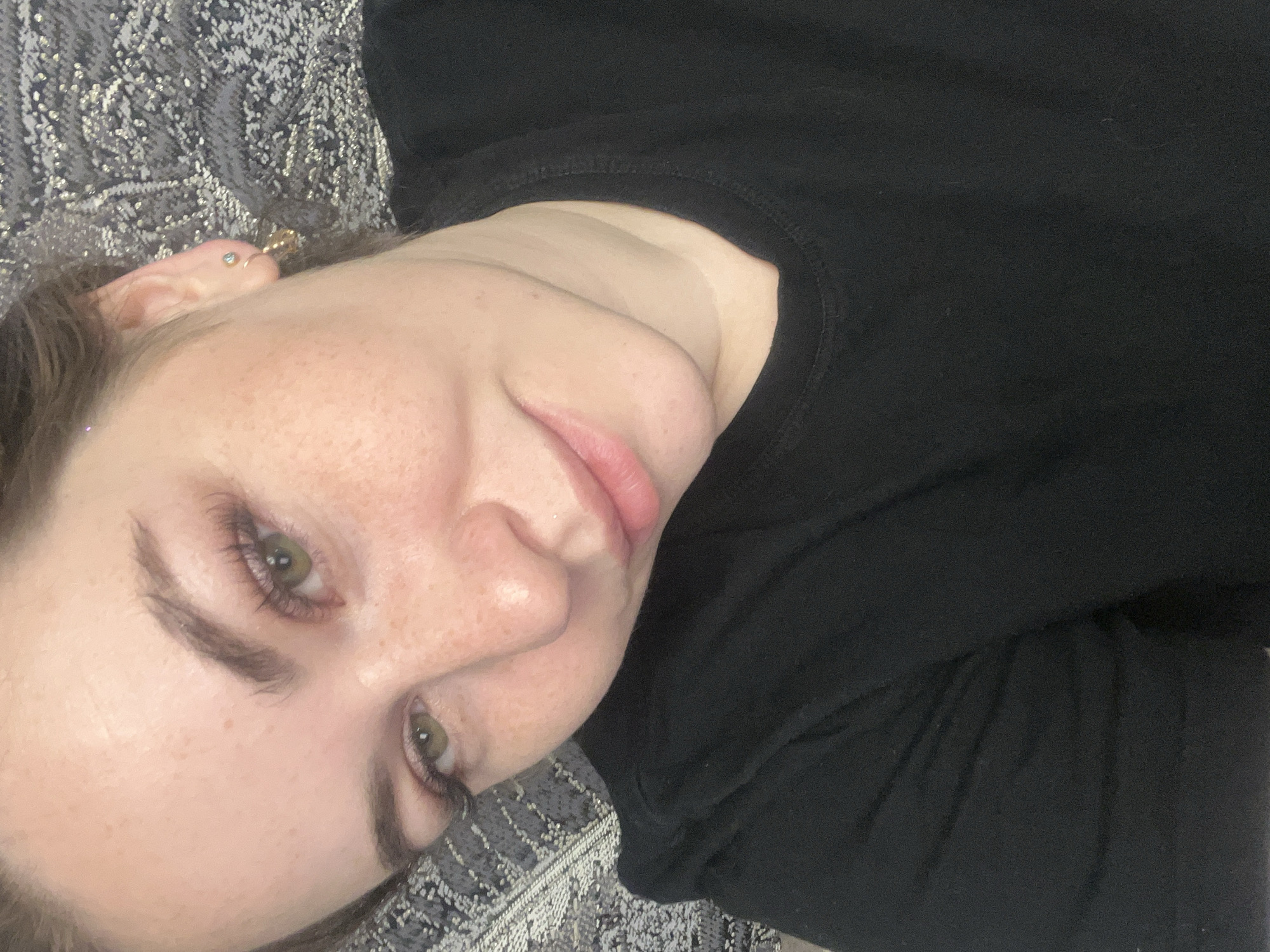 A young, light-skinned person with freckles is lying down. They have green eyes, dark eyebrows, and pink lips. They are wearing a black shirt. An ear is visible on the upper left, adorned with multiple earrings. The person is lying on a textured grey and white surface. The location is not discernible from the image.