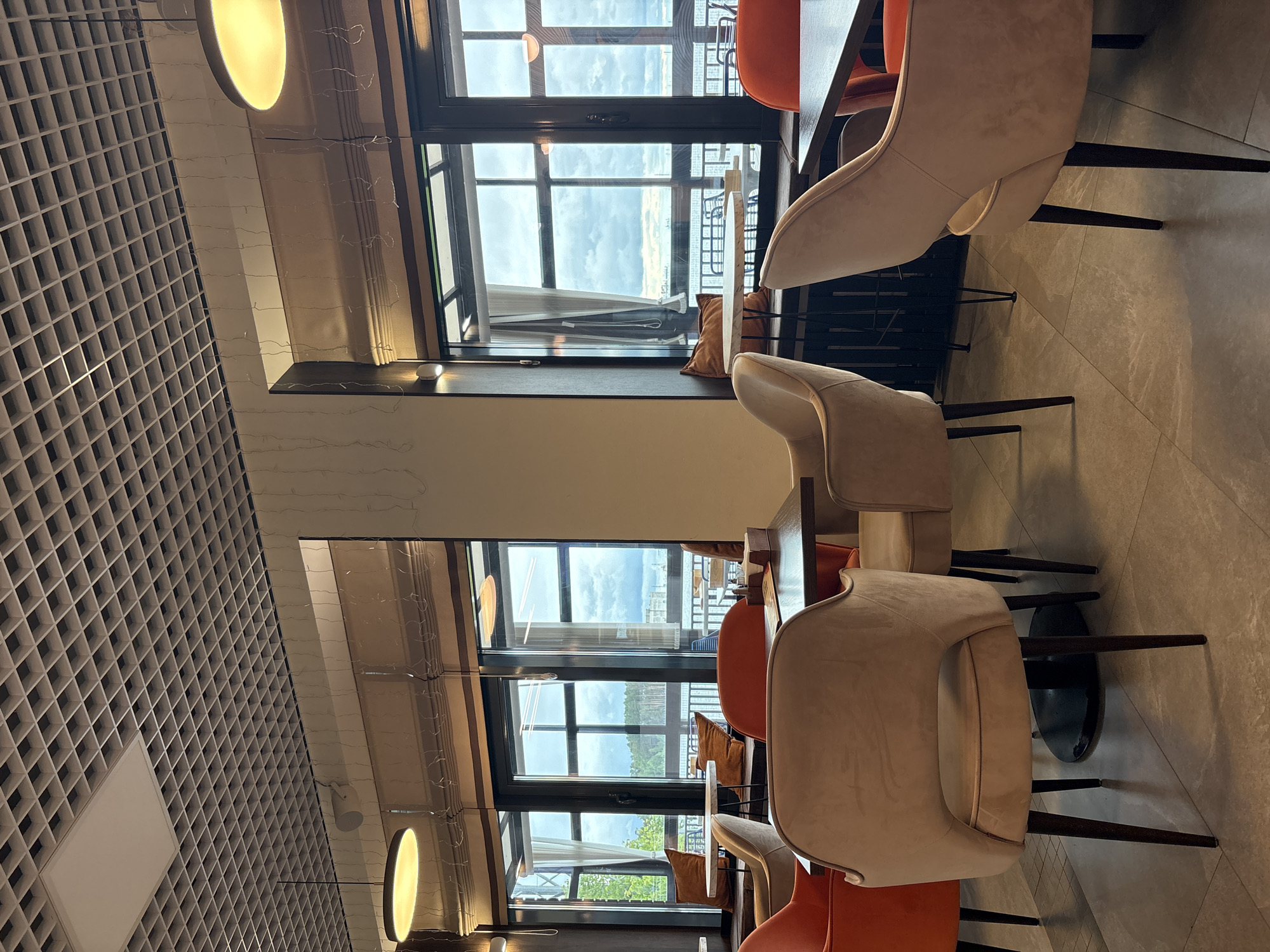 This image presents the interior of an unoccupied modern cafe or restaurant situated in the city of Chaikovsky, Russia. The setting is characterized by a prominent light-colored, grid-patterned open-cell ceiling, featuring integrated rectangular light fixtures and two visible circular pendant lights with warm yellow illumination. Numerous string lights are suspended vertically along the upper wall sections above the windows. Large, dark-framed windows dominate the background, offering a view of an overcast sky and a body of water, with a tree-lined shore visible in the distance. Brown roll-up blinds are partially lowered along the top of the windows, and a white sheer curtain is visible on the far left window panel. Wide window sills function as bench seating, adorned with orange throw pillows. The furniture includes multiple upholstered chairs with slender dark legs; some are a light beige/taupe color, while others are a vibrant orange. These chairs are arranged around dark-topped tables, some of which hold menus and condiment dispensers. The floor is covered with large, light-toned square tiles. A white circular speaker is mounted on an upper wall section to the left. No individuals are present in the scene.