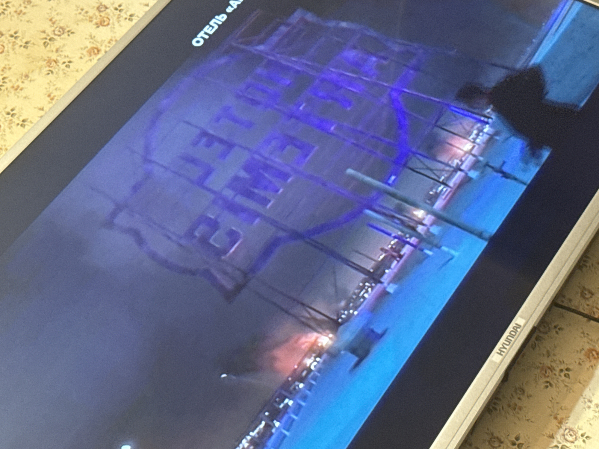 The image shows a Hyundai television screen displaying a nighttime scene. In the upper left corner of the screen, the Russian word "Отель" (Hotel) is visible. The primary subject is a large sign with the words "LET ME 2" etched on it with a blue outline. The scene is set outdoors, indicated by the lighting fixtures of the city, Mytishchi, Russia. The presence of streetlights below and structures in the distance point to a city at night. A dark, human-shaped shadow is visible in the upper right of the screen.