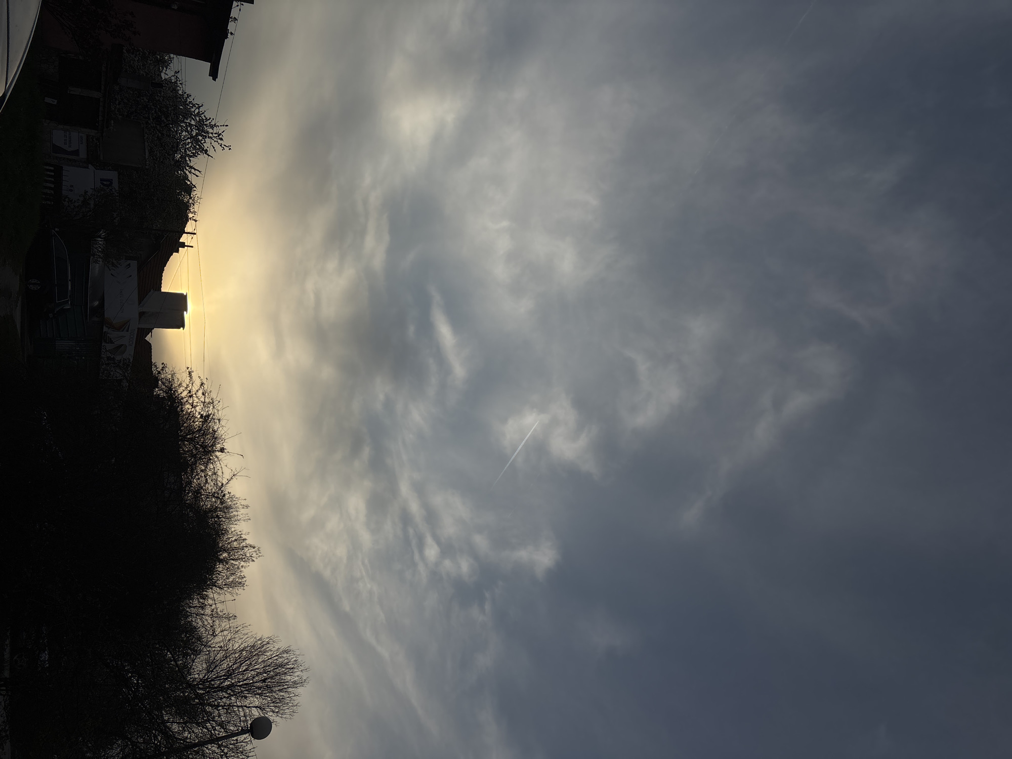 The image depicts a dusk or dawn scene in Sofia, Bulgaria, judging by the architectural style of the buildings visible on the left edge. The sun is low on the horizon, partially obscured by the silhouette of buildings and trees. The sky is filled with a mixture of gray and white clouds, with a contrail visible. A street lamp is visible in the lower left corner, obscured by tree branches.