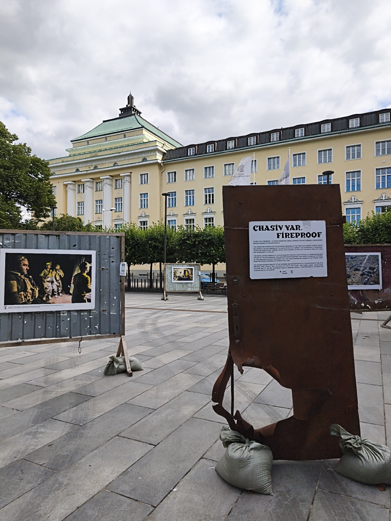 The image depicts an outdoor exhibition in Tallinn, Estonia, in front of a large, light-yellow building with a distinctive green roof and a stepped tower. In the foreground, a large, rusty, damaged metal structure, possibly a door or panel with a hole in it, stands upright. Attached to this structure is a white sign with black text that reads "CHASIV YAR. FIREPROOF." Below this title, there is descriptive text about an exhibition chronicling the defensive campaign of Chasiv Yar. Two light green sandbags are placed at the base of this metal structure for support. To the left, a grey corrugated metal panel, also on a stand, displays a framed photograph. This photograph shows several individuals in military attire. Another smaller framed photograph is visible further in the background, as is another display panel. The ground is paved with large, grey rectangular tiles. Trees with green foliage are visible on either side of the exhibition area, and a cloudy sky with patches of blue is overhead. Flags, one white with black text, are visible on poles in front of the building. The overall scene suggests a public space in a city setting, likely a plaza or square, hosting an exhibition related to military events.