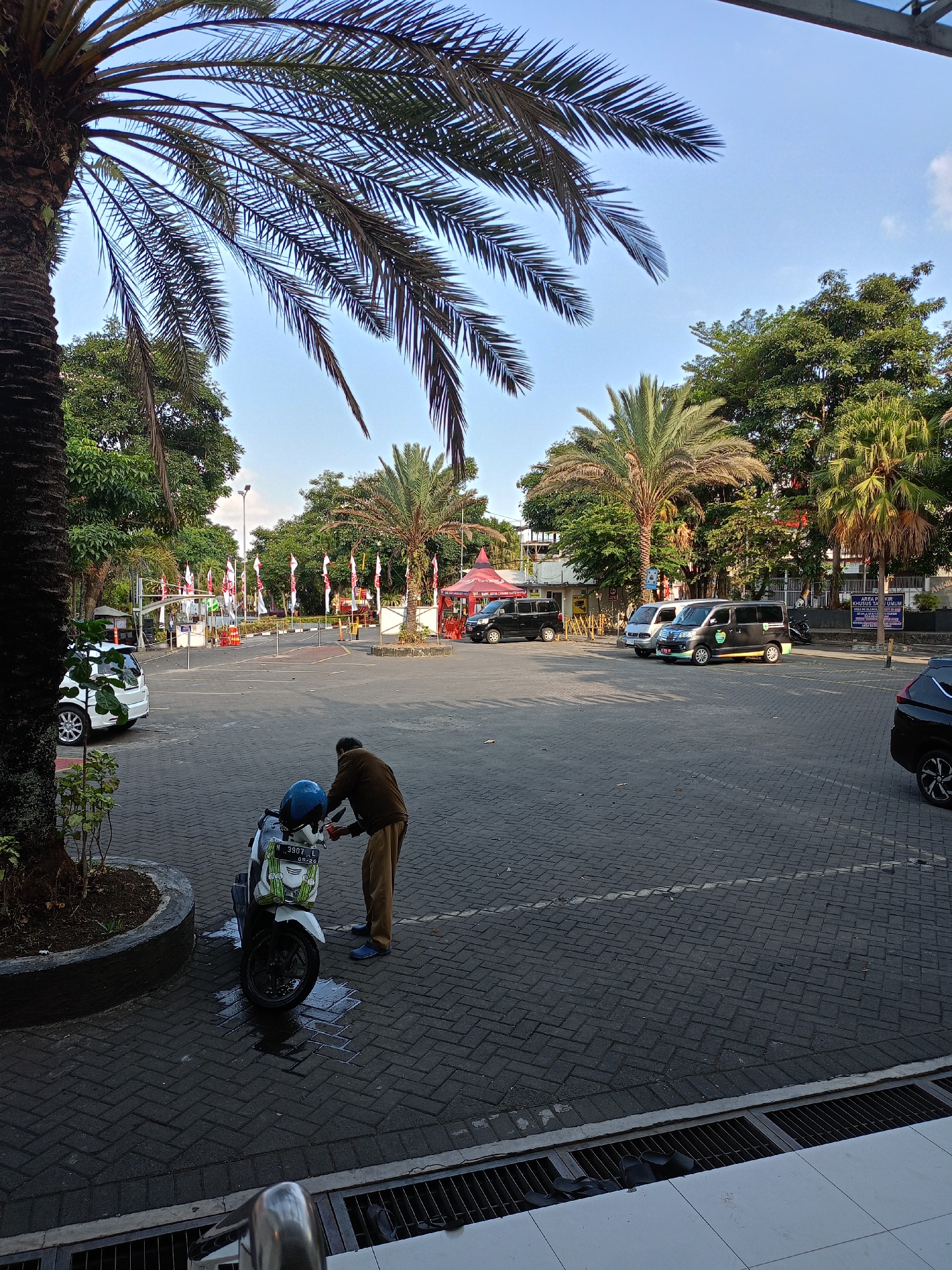 In the city of Batu, Indonesia, a person in brown clothing and blue shoes is cleaning a white scooter with a blue helmet in a paved parking lot. The scooter is parked near a large palm tree on the left. In the background, there are several vans, a white car, and a row of flags. The sky is clear and blue, with some clouds visible. The scene appears to be outdoors, possibly in a public area or a parking lot for a building.