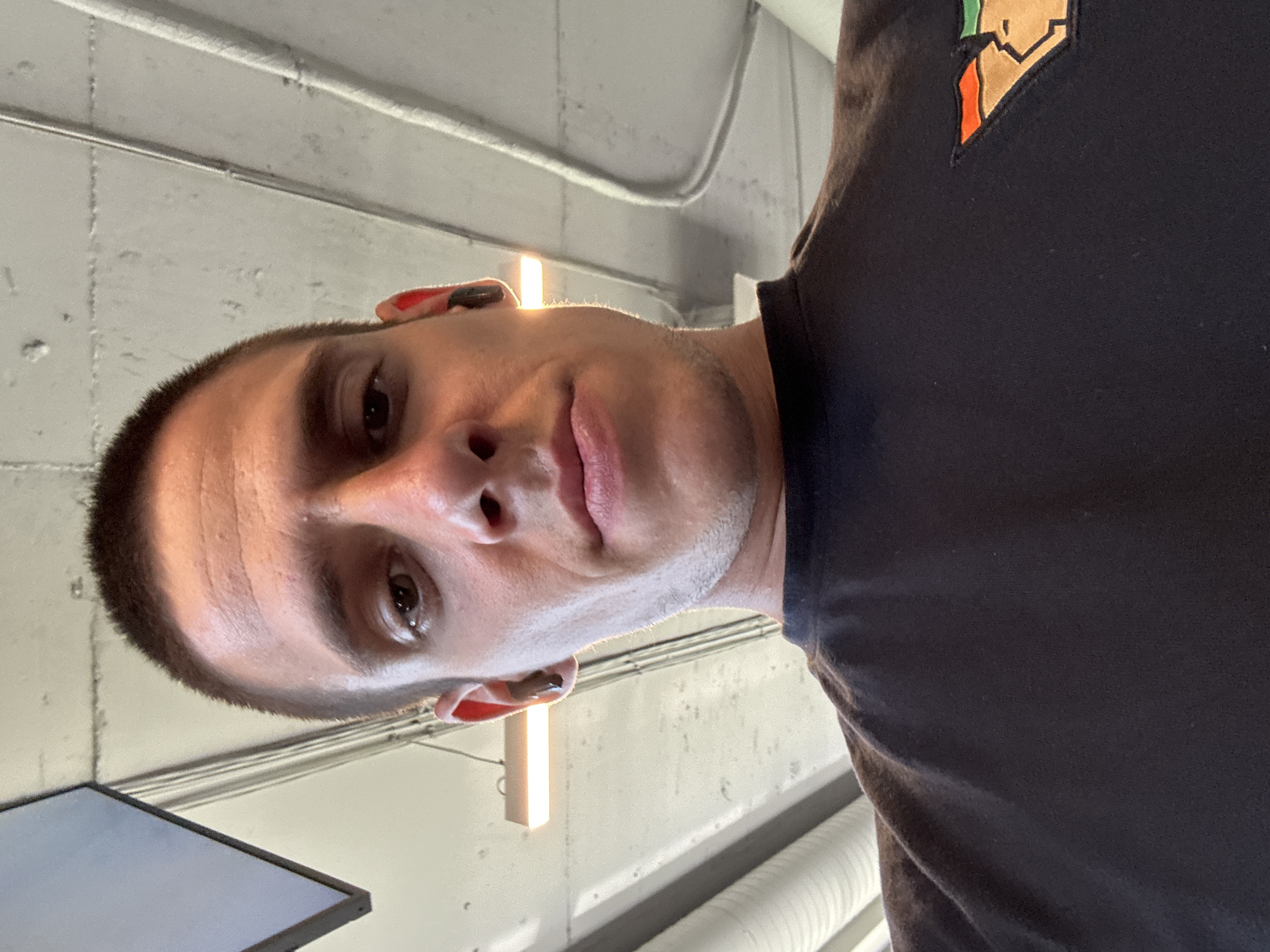 A low-angle, close-up, selfie-style photograph shows a man with a buzz cut and light stubble. He is wearing a black t-shirt with a geometric patch on the right side of his chest. The patch features angular shapes in orange, green, and tan colors, outlined in black. He has earbuds in both ears, with the right earbud appearing red. The background consists of industrial-style ceiling elements with exposed pipes and lighting fixtures, suggesting an indoor, possibly commercial or public space. One fluorescent light fixture is clearly visible. The image is well-lit, with the light source casting a slight sheen on the man's forehead and nose. No specific location in Sofia, Bulgaria can be identified from the visual elements.