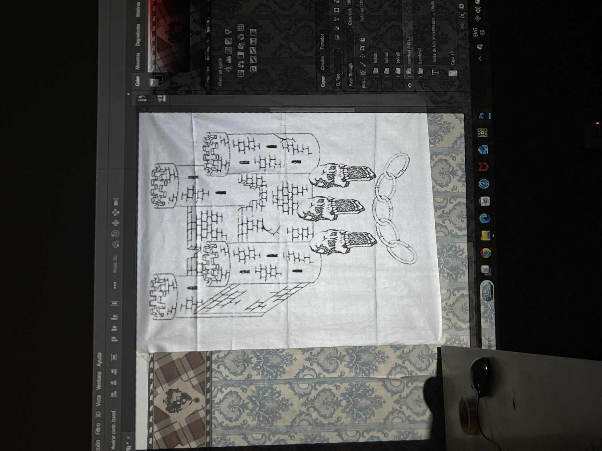 A computer monitor displays an image of a hand-drawn castle and skull design on a white fabric. The monitor also shows the Adobe Photoshop interface with various layers and settings visible in Spanish. The base of the monitor is resting on a surface with a blue and white patterned wallpaper design, and adjacent to it is a white table with a black computer mouse and a roll of tape. The image gives no information about the city of Ushuaia, Argentina.