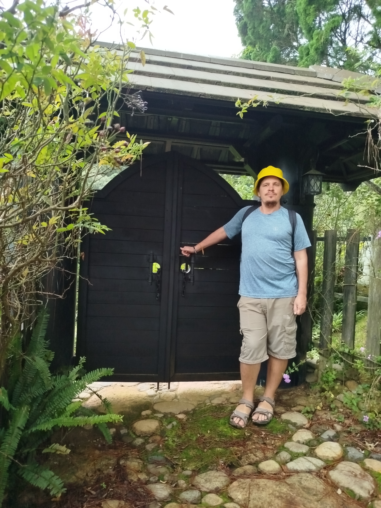A man wearing a yellow hat, blue t-shirt, and khaki shorts stands in front of a dark wooden gate with chains. He is wearing sandals. He has a backpack on his back. The gate is open, revealing a glimpse of greenery behind it. The man's left hand is on the gate handle. The ground is covered with rocks and moss. In the background, there is a wooden structure with a roof and some trees. The scene appears to be in a garden or park setting.
