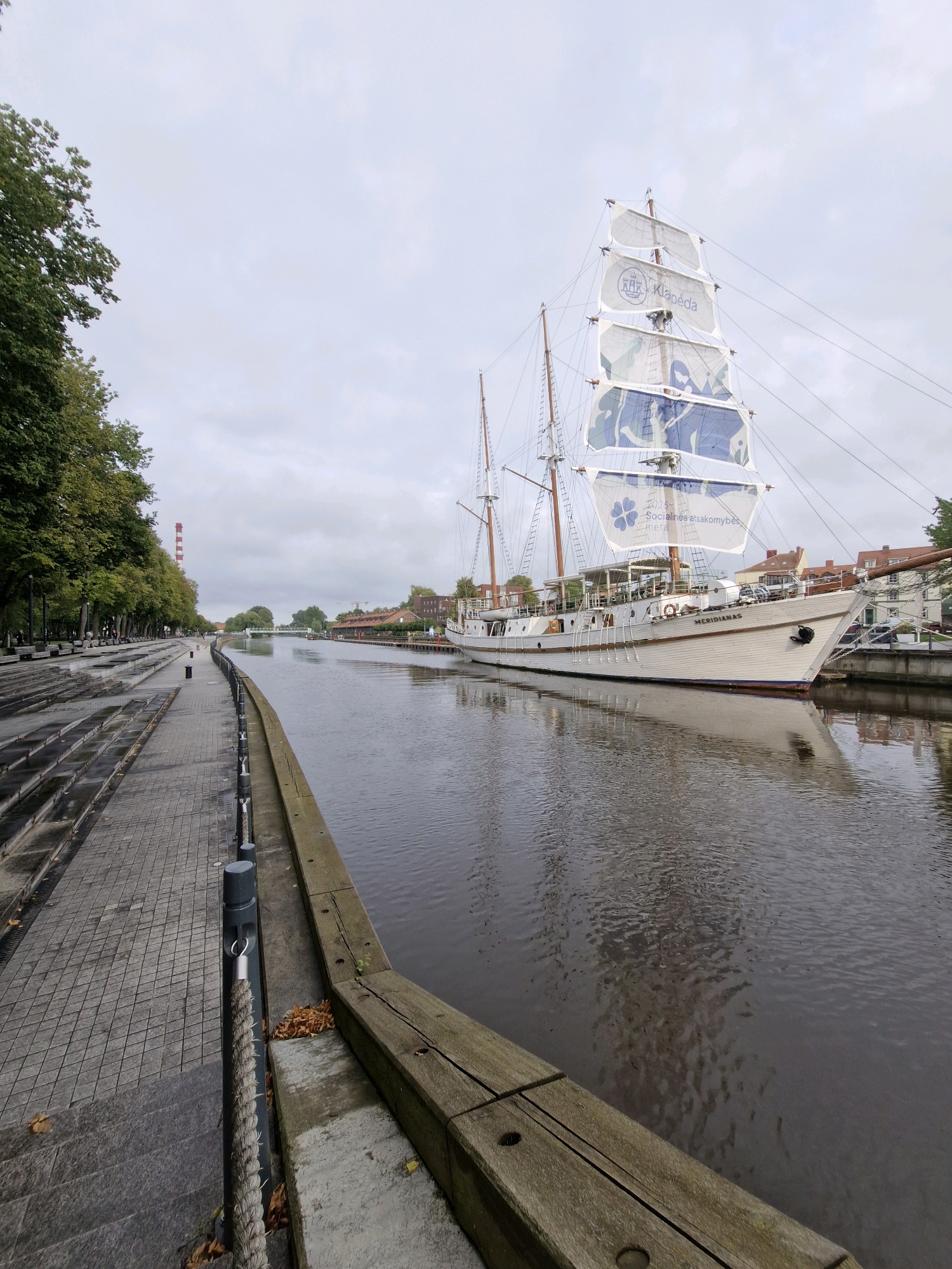 A tall ship, named "MERIDIANAS", is docked on the right side of the Danė River in Klaipėda, Lithuania. The ship is white with visible masts and sails, some of which have blue graphics and text that appears to be in Lithuanian. The river is calm and reflects the overcast sky and the ship. On the left side of the river, a paved walkway runs alongside a tree-lined embankment with benches. The walkway has a tiered design with stairs leading down to the water's edge. Several bollards and a thick rope are visible along the edge of the walkway. In the background, a bridge crosses the river, and buildings are visible on the far bank. A tall, red and white striped industrial chimney is also present in the distance. The sky is cloudy, suggesting an overcast day. No people are clearly visible in the image, though distant figures might be present on the walkway or on the ship.