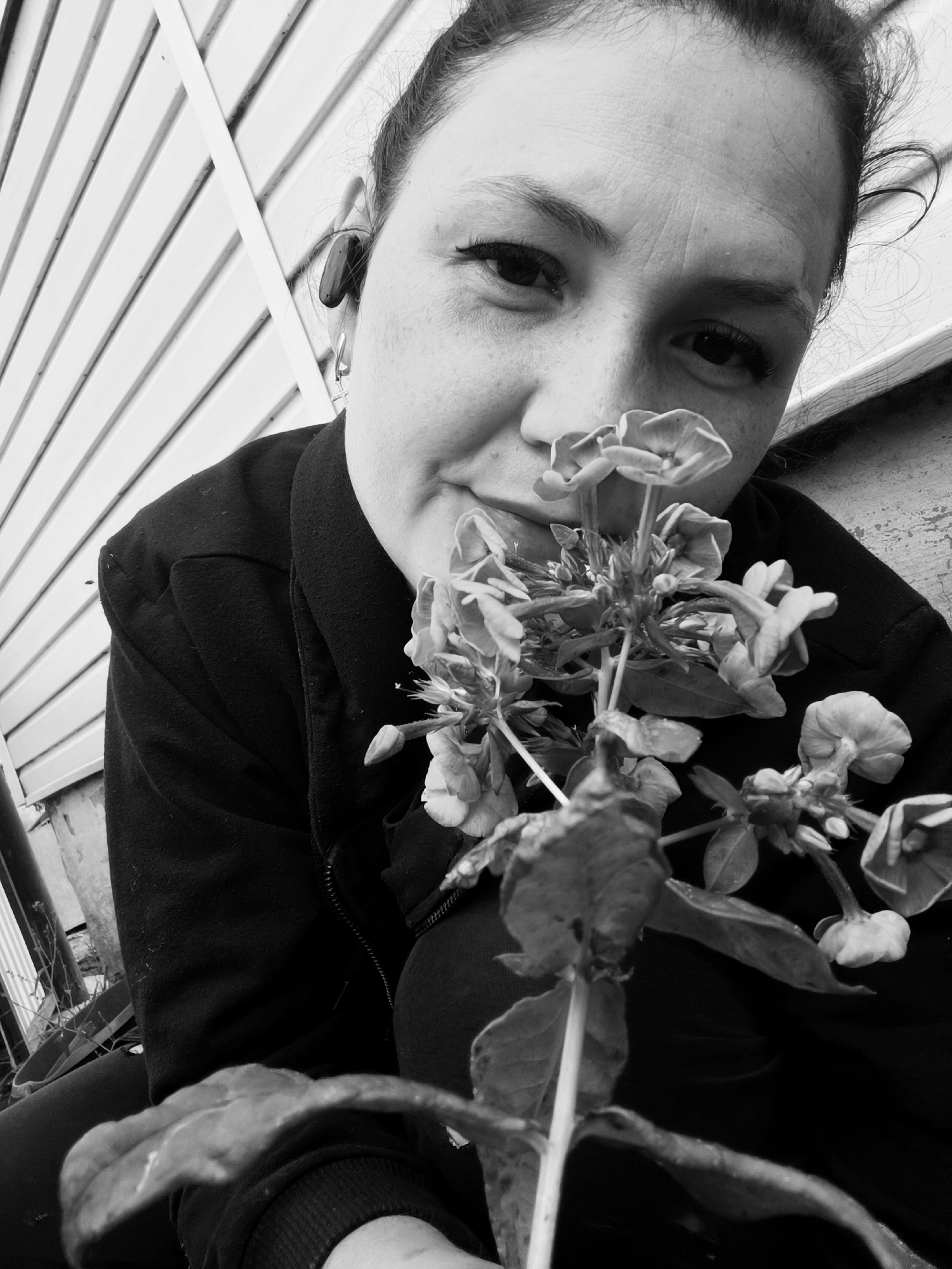 The black and white, close-up photograph shows a woman holding a flowering plant up to her face. She is wearing a dark jacket and has dark hair pulled back. An earbud is visible in her right ear. The background consists of a striped siding and a weathered wooden surface, suggesting an outdoor setting such as a residential area. There is no indication of the city of Karsha, Russia, or any specific urban environment. The image focuses on the woman's face and the plant, implying an interaction between the two. The overall mood is contemplative.