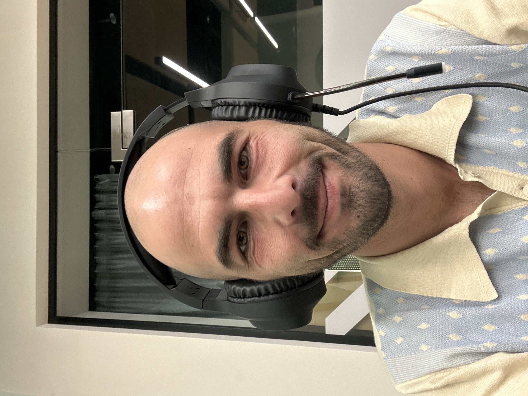 A close-up, indoor shot depicts a man wearing black headphones with a microphone. He has a shaved head, light skin, and a trimmed beard. He is wearing a light blue and tan collared shirt. The background shows a window or pane of glass with horizontal light fixtures reflected on it. The image does not provide sufficient information to determine a specific location in Budva, Montenegro, or to describe any actions or interactions.