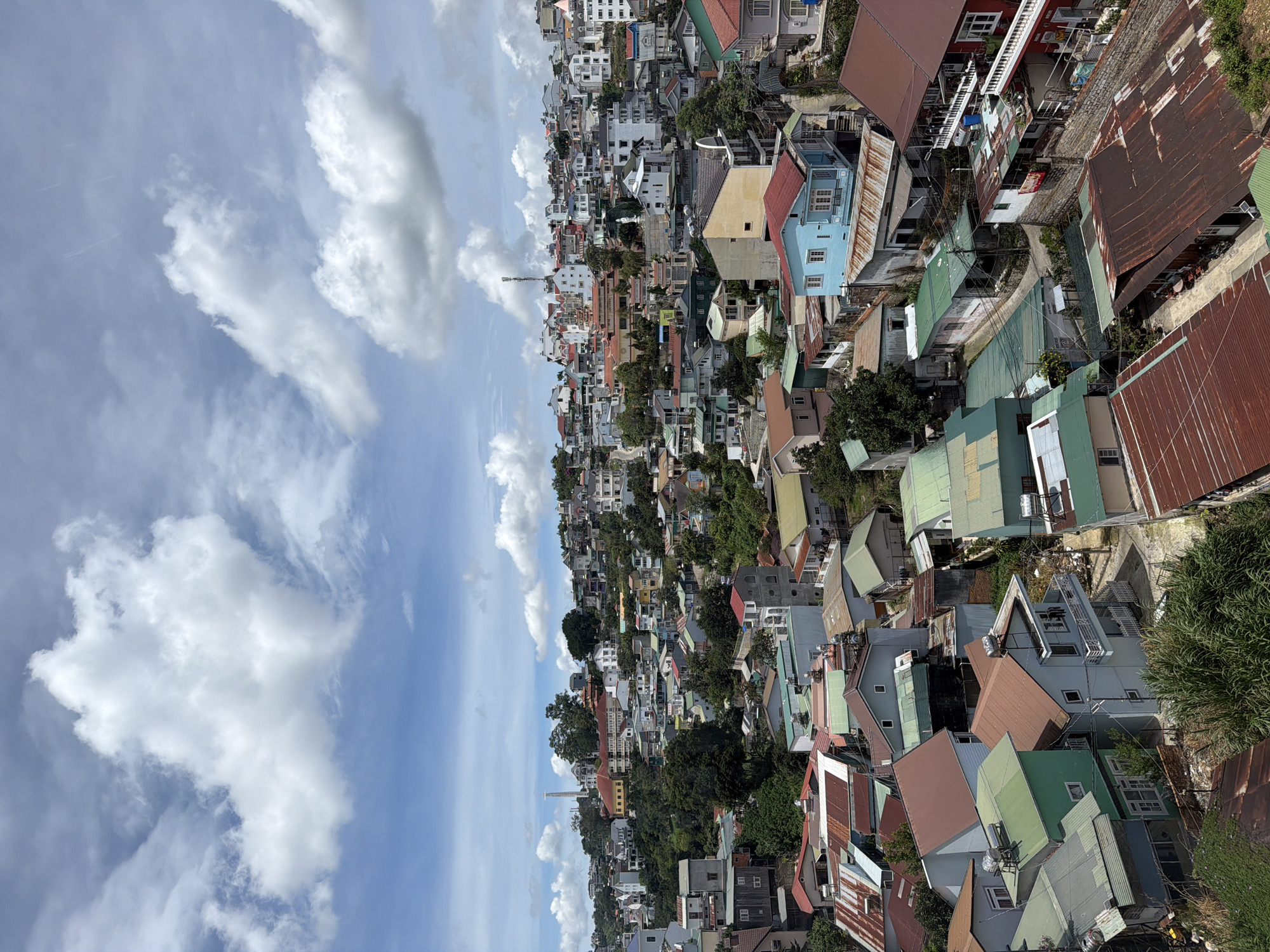 The image shows a panoramic view of a hillside residential area in Da Lat, Vietnam, under a partly cloudy sky. The houses are densely packed, with a variety of architectural styles and roof colors, including red, green, and brown. Many of the roofs are made of corrugated metal, some showing signs of rust. The terrain is uneven, with buildings appearing at different levels on the slope. Lush green trees are interspersed among the houses, and some areas are overgrown with vegetation. In the distance, taller buildings and a tower are visible on the highest part of the hill. The scene is captured from an elevated vantage point, offering a broad perspective of the urban landscape and its natural surroundings. There are no visible people or specific actions taking place.