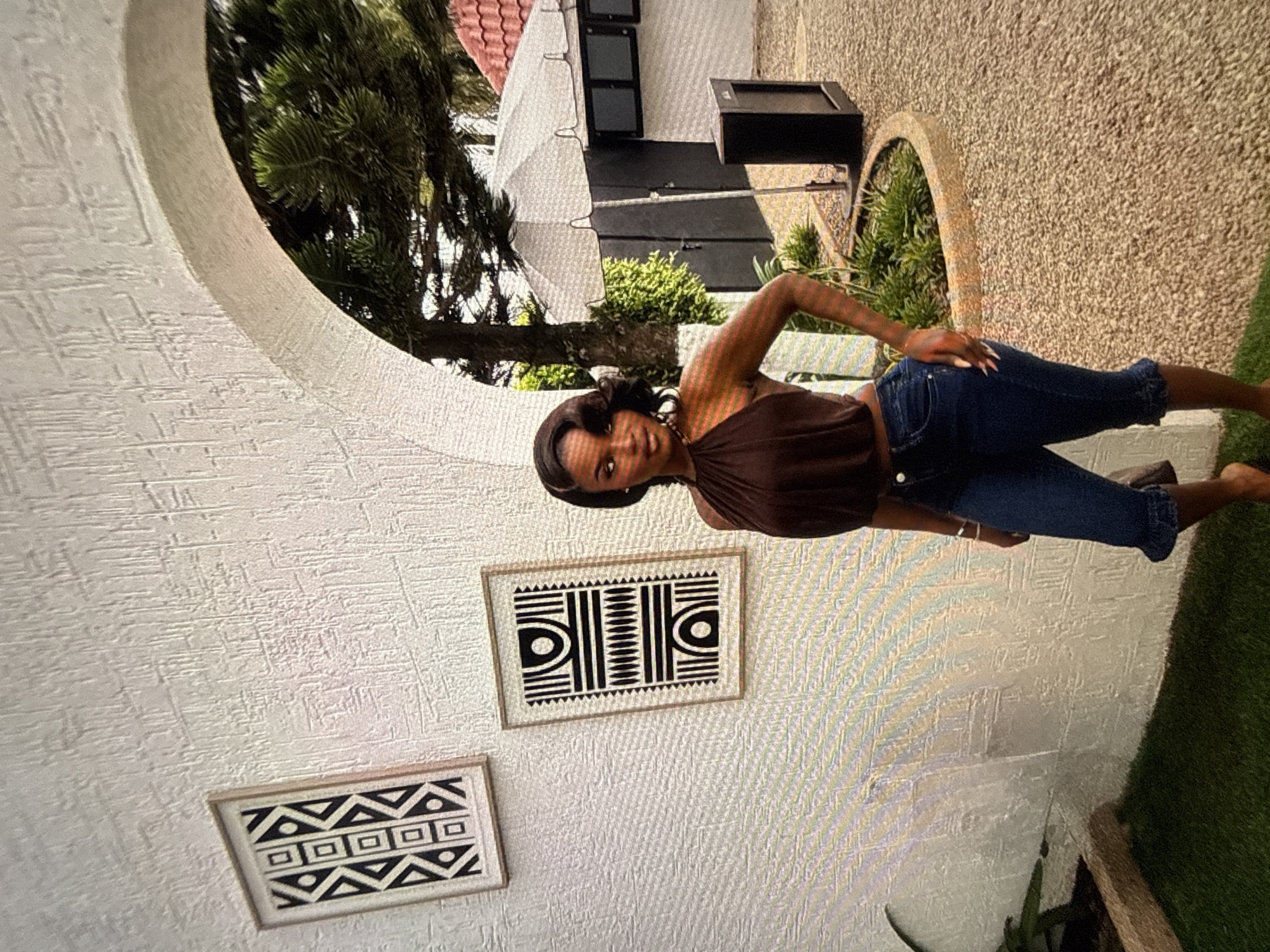 A young Black woman stands in front of a textured white wall in an outdoor setting in Abuja, Nigeria. She is wearing a brown halter top and dark blue capri jeans with frayed hems. The wall has two framed pieces of art with geometric black and white patterns. To her right, an arched walkway reveals a glimpse of trees and a white building with a red tiled roof, suggesting a courtyard or garden area. The ground is covered in gravel and some patches of green grass. A black speaker sits on the gravel near the building. The woman is looking towards the viewer with a slight smile.