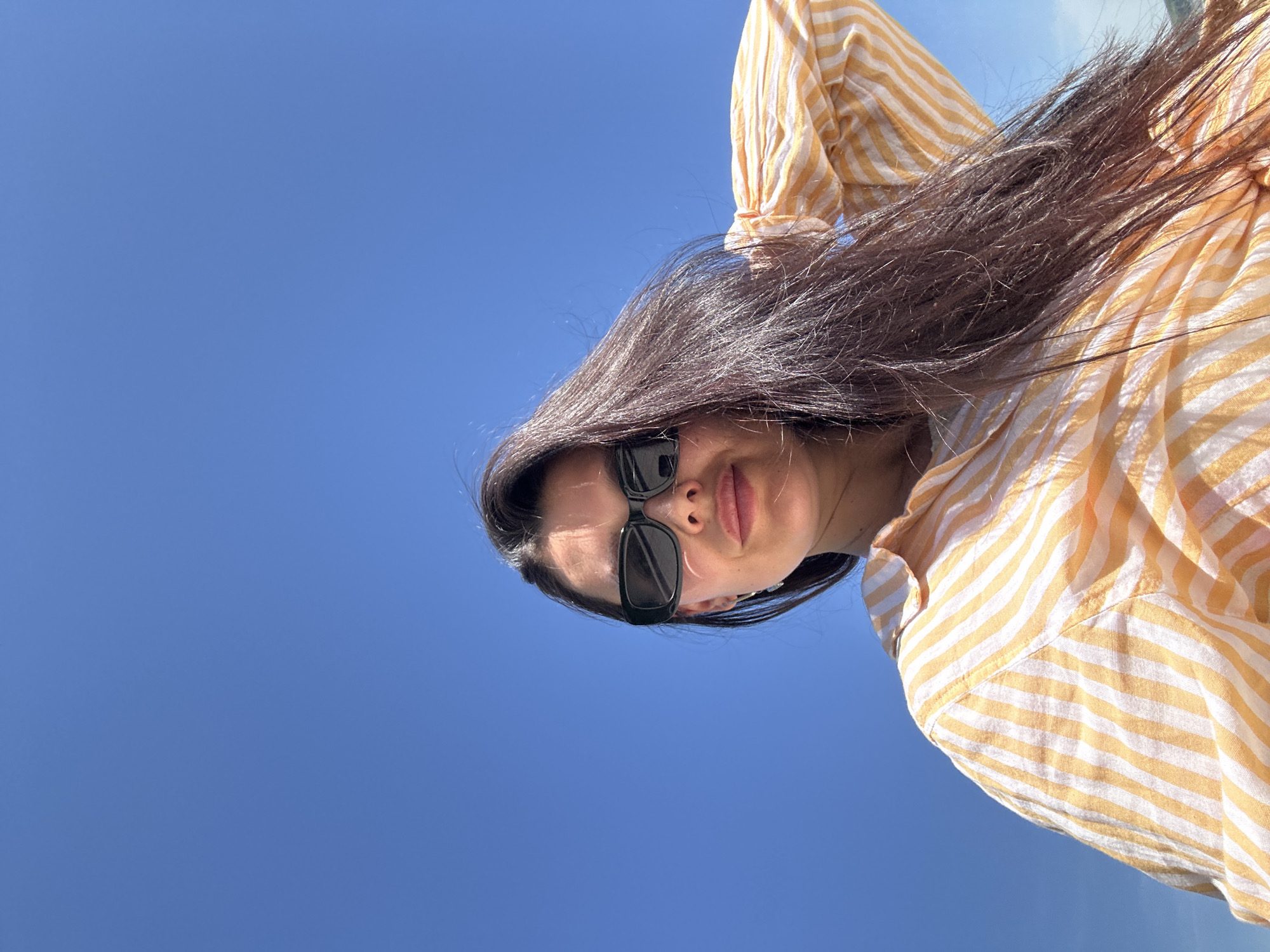 A low-angle shot depicts a woman against a clear blue sky. She is wearing sunglasses and a yellow-and-white striped shirt. Her dark hair is partially obscuring her face. The location is unknown, and there is no evidence to suggest it is in the city of Chaikovsky, Russia.