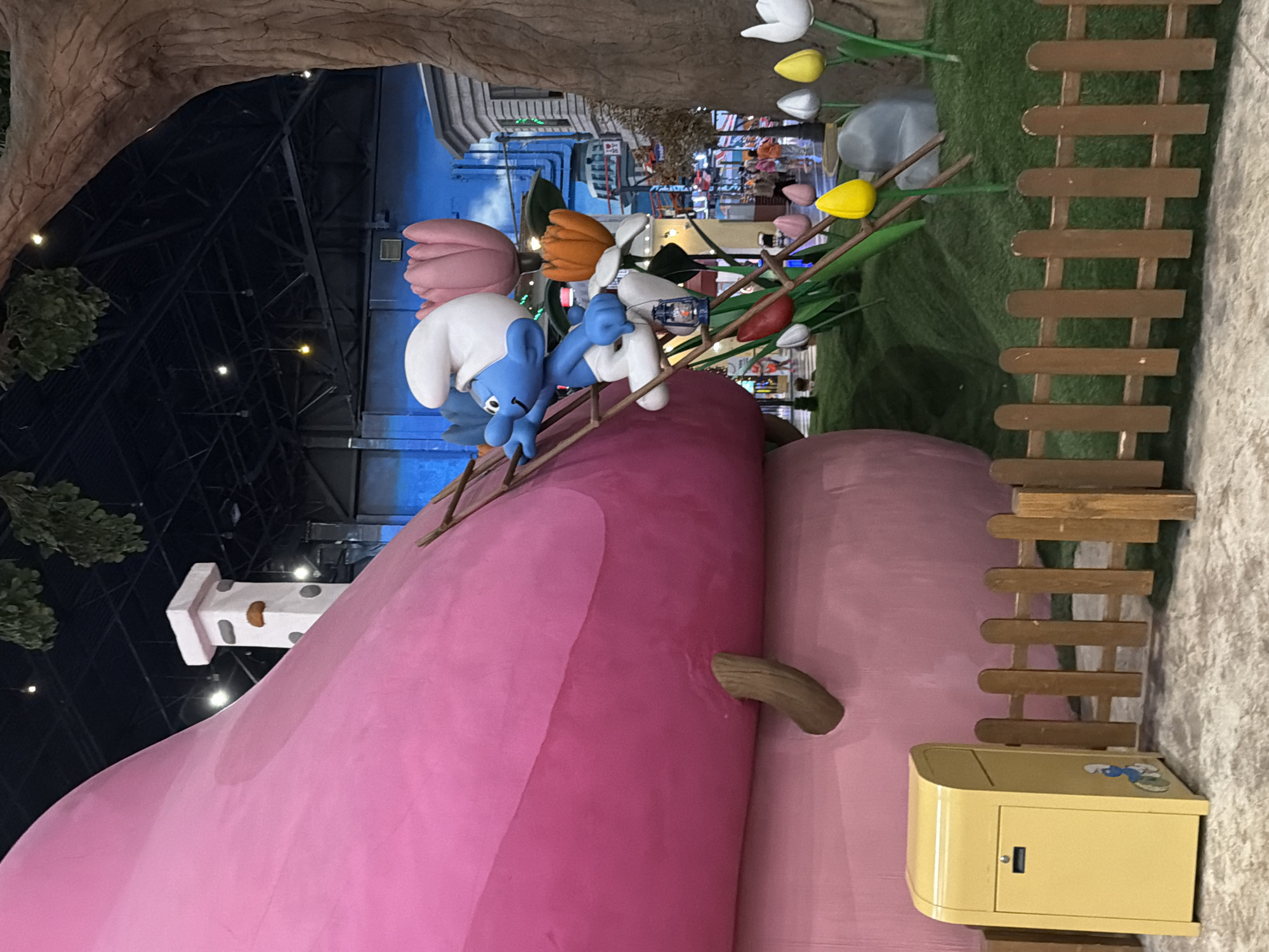 The image captures a whimsical, themed indoor environment, possibly an entertainment venue or attraction, with a strong Smurf theme. In the foreground, a bright yellow mailbox with a Smurf character graphic is visible next to a rustic wooden fence. Behind the fence, artificial grass covers the ground, dotted with large, stylized flowers in various colors, including red, orange, pink, yellow, and white. A large, pink mushroom-shaped structure dominates the left side of the frame, with a wooden ladder leaning against it. A blue Smurf character is climbing this ladder, holding a small blue lantern. To the right of the mushroom, more stylized flowers and part of a large tree trunk are present. In the background, a cityscape with buildings is depicted on a wall mural. Several blurred figures of people can be seen in the distance, suggesting a public space. The ceiling of the venue, with exposed industrial beams and lighting fixtures, is visible overhead. The overall scene evokes a playful and imaginative atmosphere.