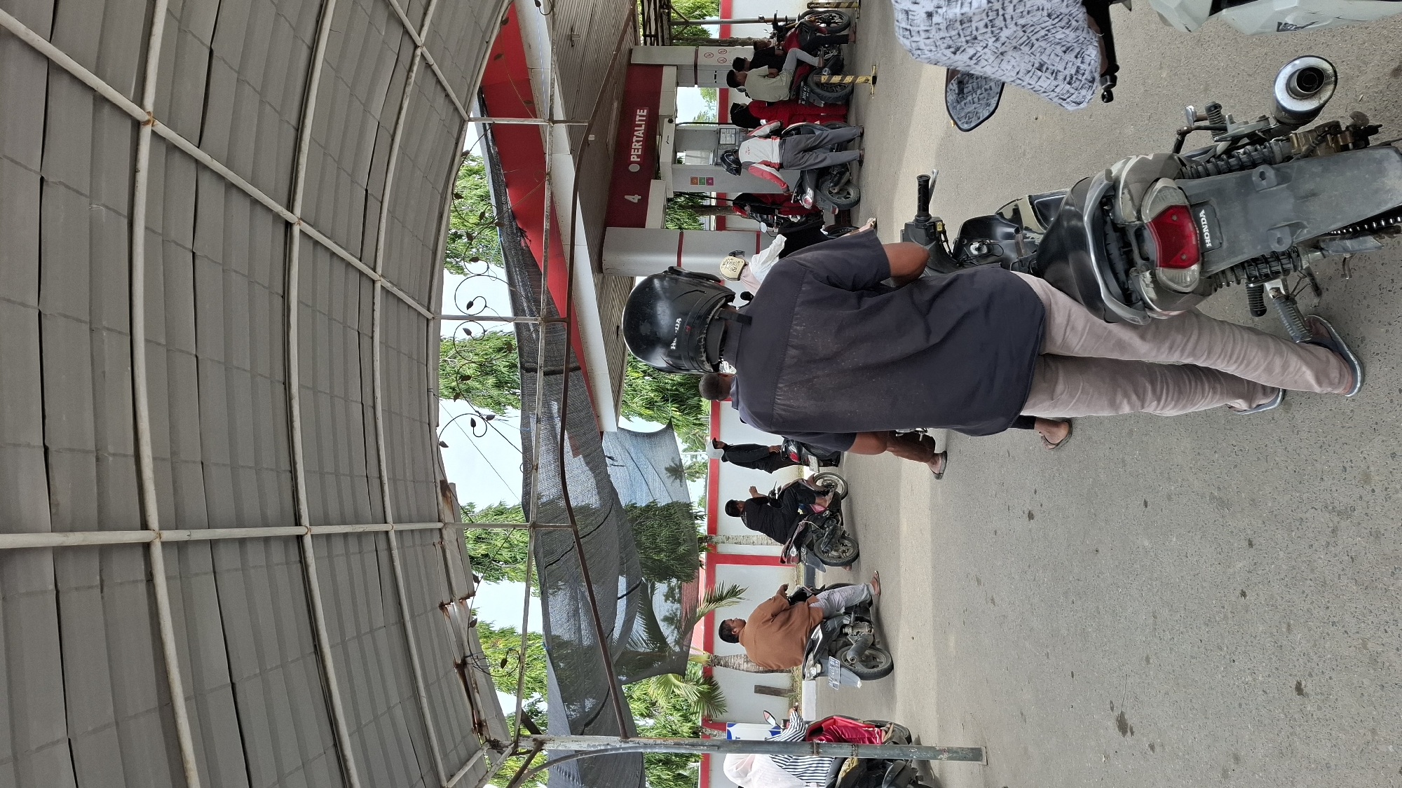 A queue of motorcyclists waits at a Pertamina gas station in Keudebayu, Indonesia. The station features a red and white canopy structure supported by metal poles. Several people are seated on or standing near their motorcycles. In the foreground, a person wearing a black shirt and black helmet is on a motorcycle, facing away from the camera. Behind them, other individuals and motorcycles are visible. The ground is paved asphalt, and trees are present in the background, suggesting an outdoor setting under a bright sky. The Pertalite branding is visible on a pump.