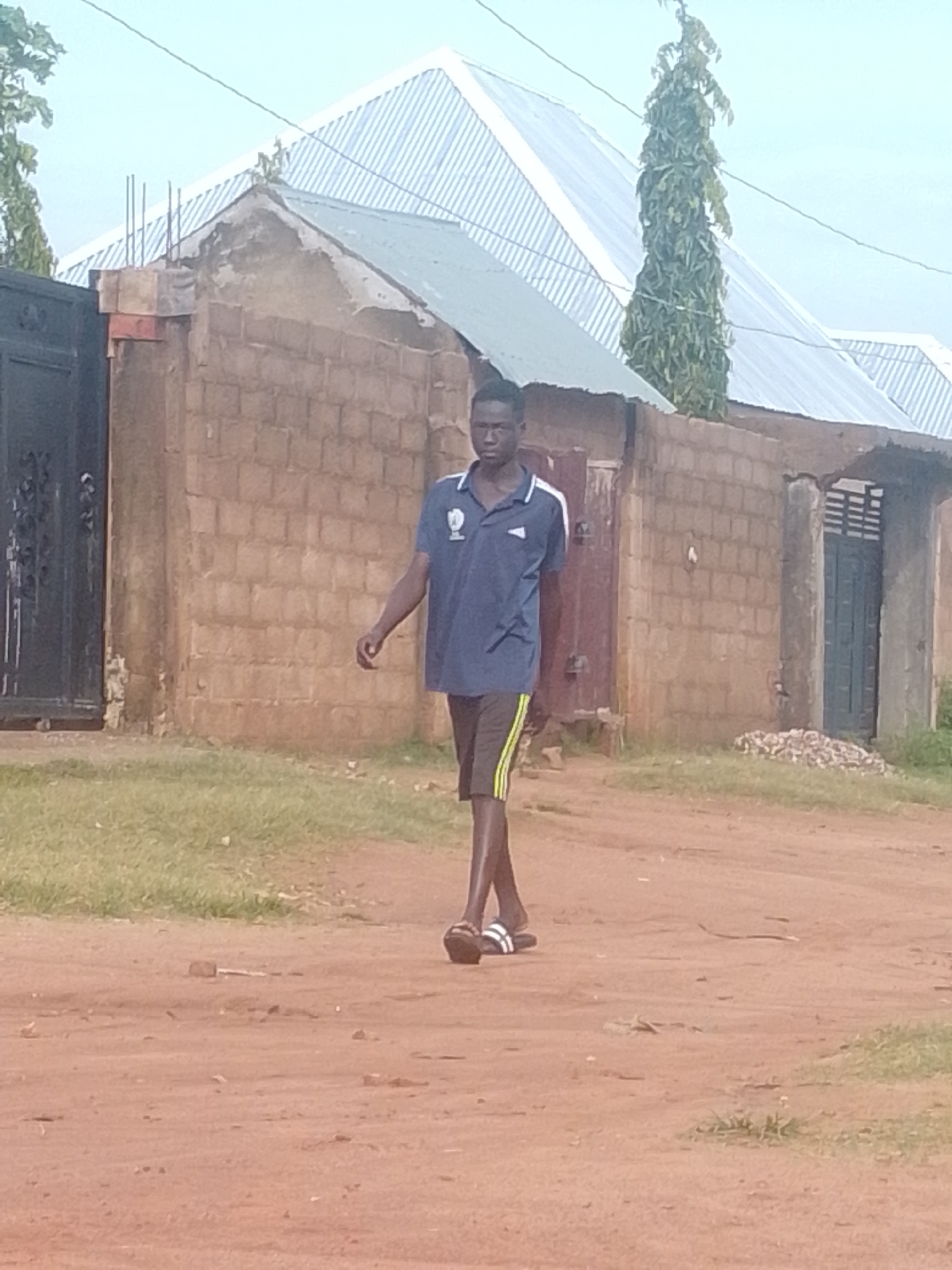 A young Black male walks on a dirt road in front of a brick building with a metal gate. The man wears a dark blue polo shirt with white accents and dark shorts with yellow stripes. He has on white sandals. The building has a light blue corrugated metal roof and multiple doors. A tall, thin tree is visible behind the building. The sky is overcast.