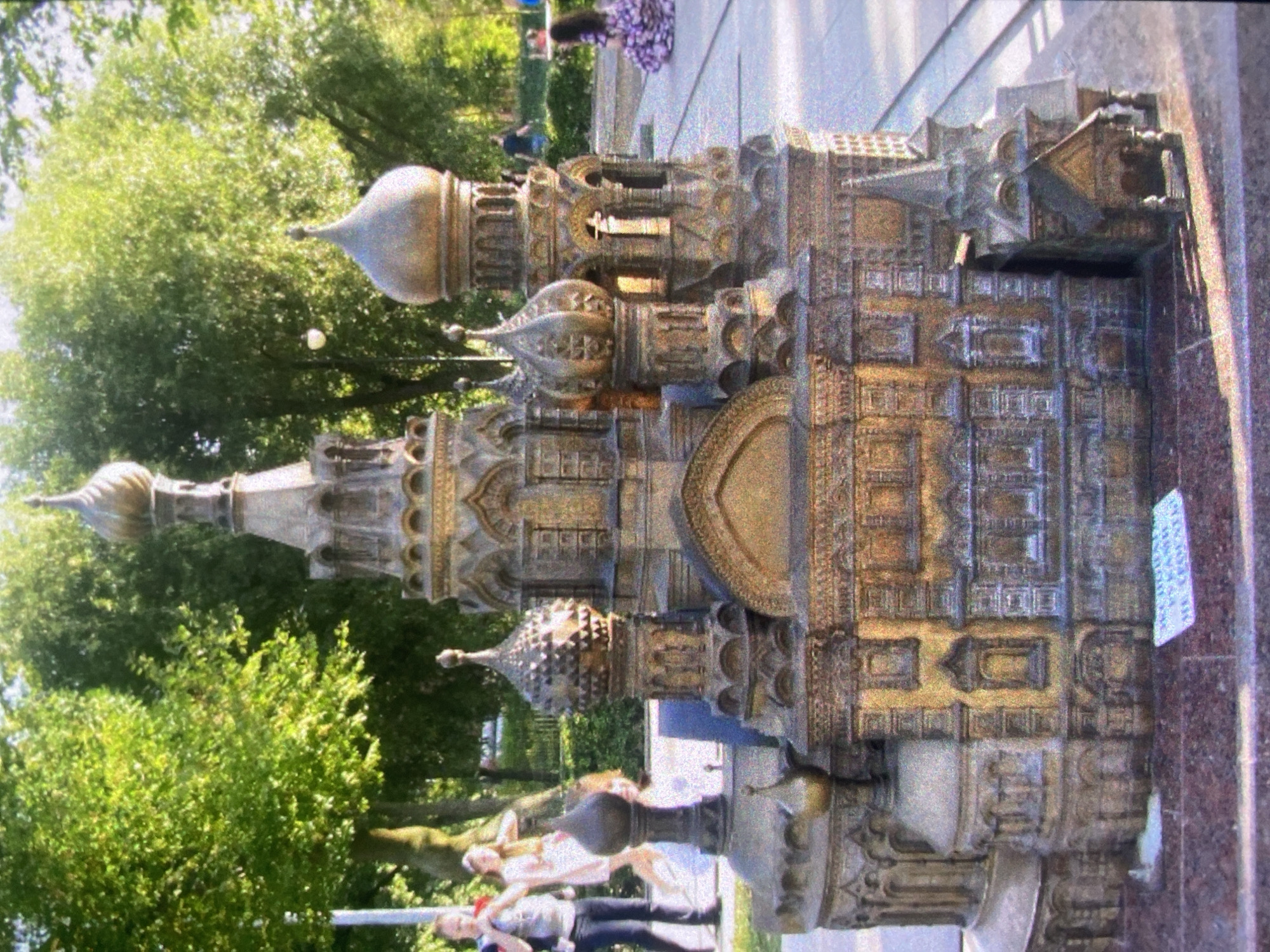 Here is a detailed description of the image:
**Overall Impression:** The image appears to be a photograph taken outdoors in a park-like setting. The primary subject is a detailed architectural model of what appears to be Saint Basil's Cathedral.
**Subjects & Objects:**
* **Architectural Model:** A detailed model of Saint Basil's Cathedral is prominent. It has multiple domes, intricate architectural details, and a bronze or gold-colored finish.
* **People:** There are at least three people visible in the foreground. They seem to be walking or standing near the model.
* **Environment:** The background includes greenery, suggesting a park or garden. There are trees and foliage visible. The model is set upon a pedestal or base, which appears to be made of marble.
**Actions & Interactions:** The people are simply present in the scene.
**Location:** The architectural model of Saint Basil's Cathedral suggests the location is in Saint Petersburg, Russia.