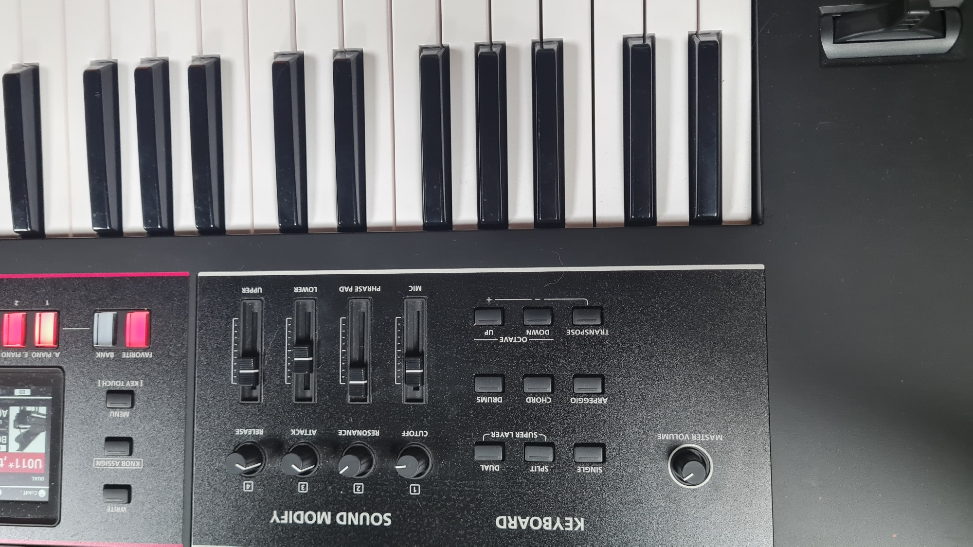 The image shows a close-up of a keyboard, likely an electronic synthesizer or digital piano, with a black control panel.
The keyboard's keys are visible at the top of the frame.
The control panel features several knobs, sliders, and buttons, labeled with functions such as "MASTER VOLUME," "SOUND MODIFY," "KEYBOARD," "OCTAVE," "TRANSPOSE," "MIC," "PHRASE PAD," "LOWER," and "UPPER."
Buttons for "ARPEGGIO," "CHORD," "DRUMS," "SUPER LAYER," "DUAL," "SPLIT," and "SINGLE" are present.
Sliders control "UPPER," "LOWER," "PHRASE PAD," and "MIC" levels.
Knobs adjust "CUTOFF," "RESONANCE," "ATTACK," and "RELEASE."
Buttons with the labels "WRITE," "KNOB ASSIGN," "MENU" and "KEY TOUCH" are shown.
The buttons "A. PIANO E. PIANO" and "FAVORITE BANK" are illuminated red.
A display screen is shown on the left with the text "DUAL".
A possible location of Nahariyya, Israel cannot be determined from the information.