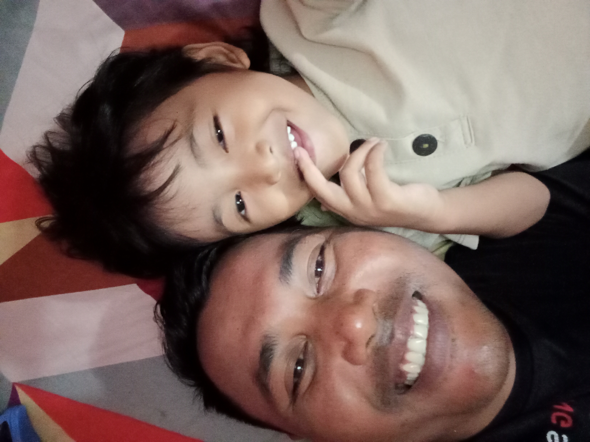 In a selfie taken indoors, a young child with dark hair lies with their head resting on an adult male's chest. The child, positioned above the adult, is looking towards the camera with a playful expression, touching their lips with a finger and showing their teeth. The child is wearing a light-colored shirt with a dark button. The adult, whose face is in the foreground and upside down relative to the child, is smiling broadly, revealing white teeth. The background consists of a colorful patterned surface, possibly a bedspread or mat, with geometric shapes in shades of red, brown, purple, and white. The scene appears to be a casual moment captured in Bloro, Indonesia.