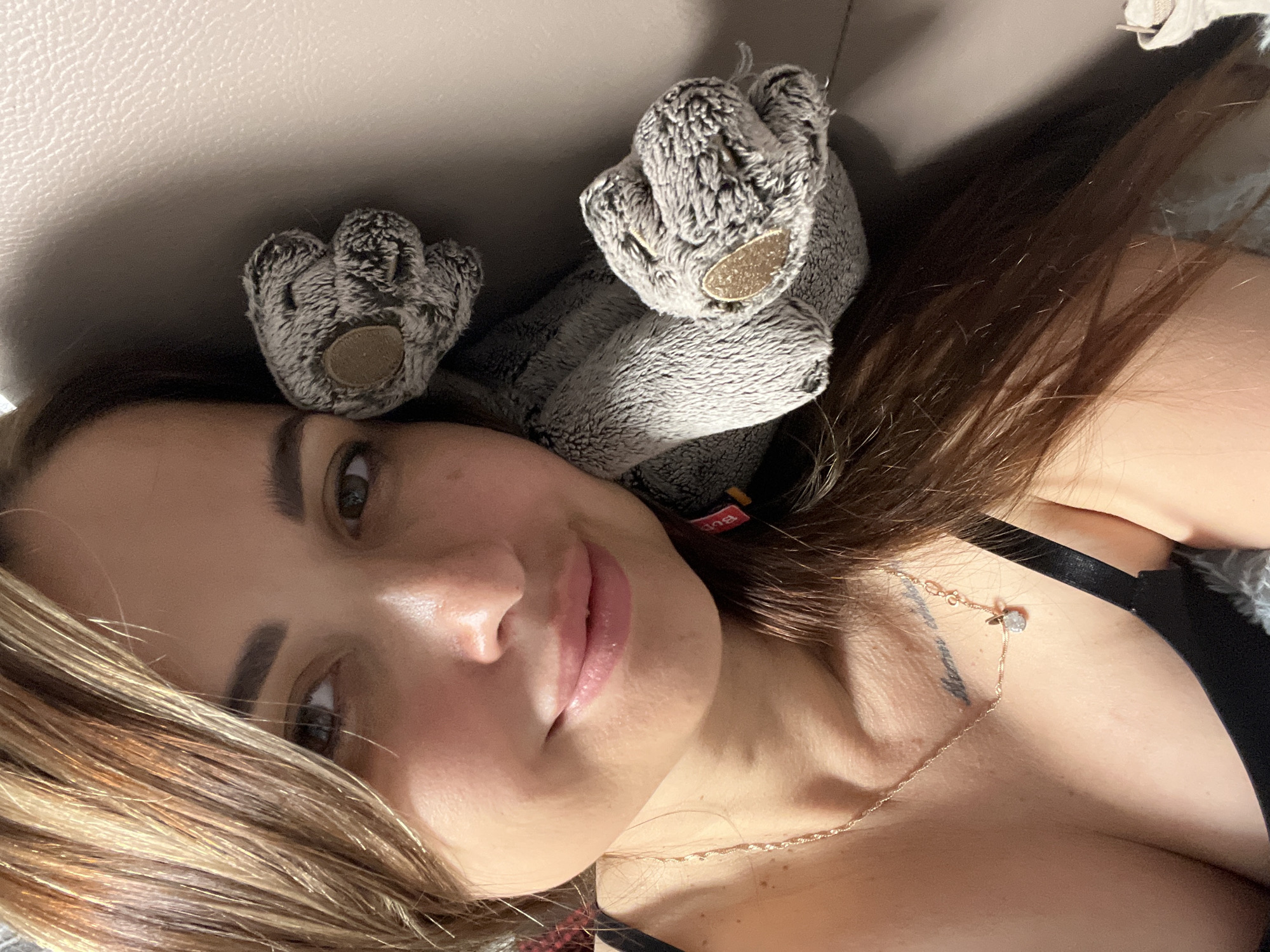 A selfie of a woman is captured from a high angle, her head resting against a textured, light-colored surface, possibly a couch. She has long, brown hair with blonde highlights, framing her face. Her eyes are a light brown, and she wears light makeup. Her lips are slightly parted in a gentle smile. She is wearing a black bra and a delicate gold necklace with a small pendant. A tattoo is visible on her collarbone. To her right, a plush grey stuffed animal, possibly a bear paw, is tucked beside her head. The background is a muted, textured surface, suggesting an indoor setting.