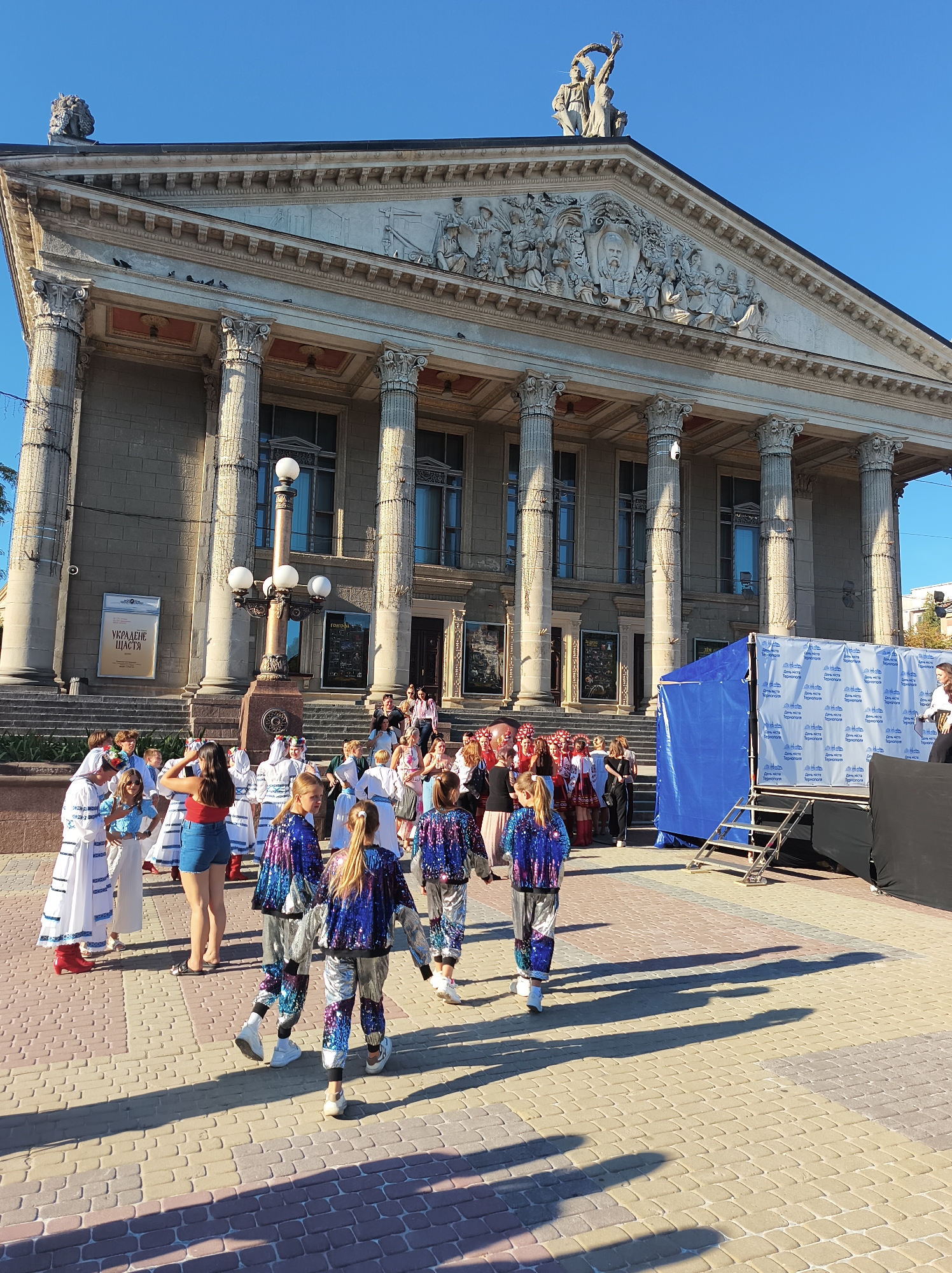 In front of a grand stone building with classical columns, a group of children in colorful, sequined outfits and traditional Ukrainian attire are gathered. Some are walking, while others stand and converse. The building appears to be a public institution, possibly a theater or a government building, with a large pediment featuring sculptures. A blue tent structure with a logo is set up to the right, suggesting an outdoor event or performance. The ground is paved with interlocking bricks, and long shadows stretch across the scene, indicating it's a sunny day. The overall atmosphere suggests a cultural gathering or a performance in Ternopil, Ukraine.