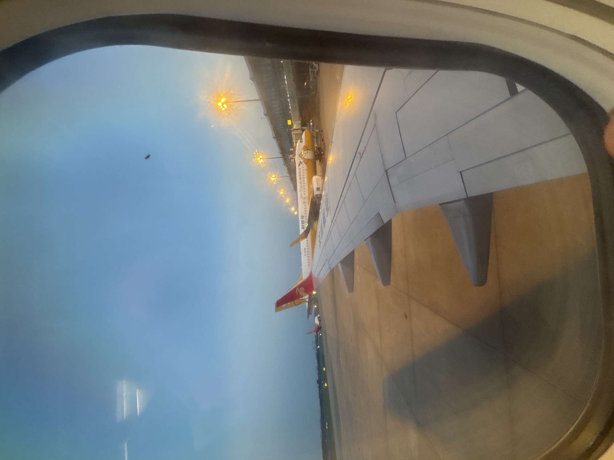 The image is taken from an airplane window, looking out at an airport tarmac during dusk. The sky is a gradient of blue and gray, with the lights of the airport beginning to illuminate the scene. In the foreground, a portion of an airplane wing is visible, with its characteristic metallic panels and rivets.
Further out on the tarmac, several airplanes are parked. The most prominent is a yellow airplane with a cartoon duck logo on its side and the words "Air Travel" and Chinese characters on its fuselage. This plane is connected to a jet bridge. Another airplane with a red and gold tail fin is visible in the middle ground. In the background, airport terminal buildings and rows of illuminated light poles are present, casting a warm glow. The tarmac itself is paved and appears to be marked with lines. The overall impression is that of an airport at a transitional time of day, with planes preparing for or having just completed flights. The location is identified as Xinwu District, China.