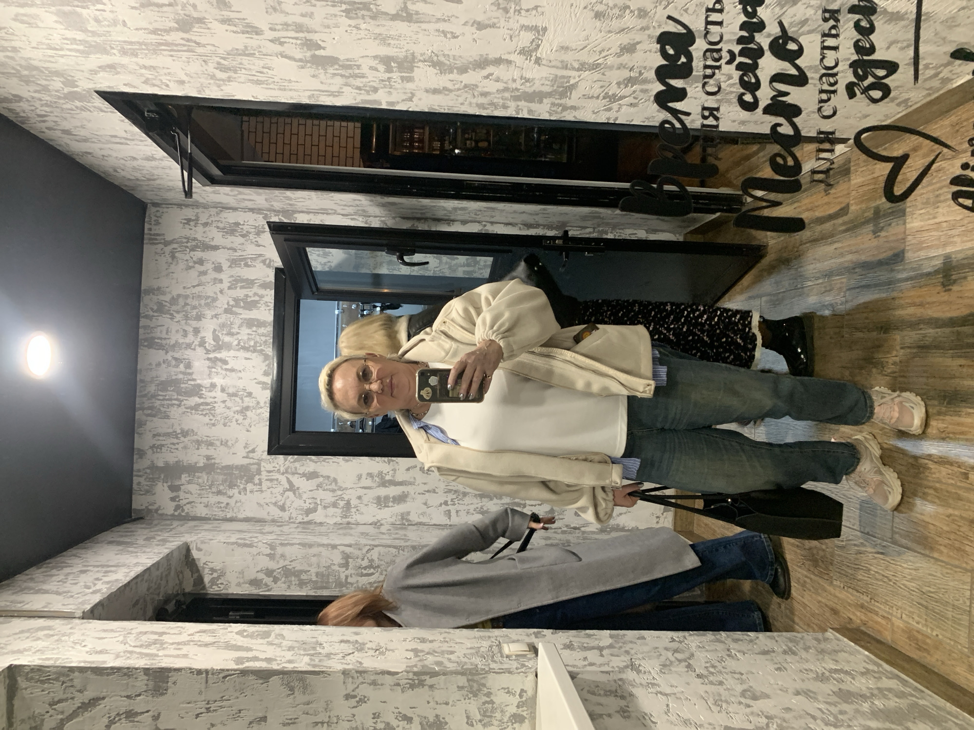 An indoor, vertically-oriented selfie taken in a mirror features a blonde woman with glasses, wearing a white top, a light beige jacket, blue jeans, and light-colored sneakers. She holds a smartphone to capture her reflection, with a black handbag hanging from her arm. Her expression is neutral as she focuses on the phone.
Behind the main subject, another person with brown hair, dressed in a grey coat and dark jeans, is partially visible in the mirror's reflection. The setting appears to be a hallway or changing room, characterized by textured, grey and white patterned walls that resemble distressed wallpaper or paint. The floor is made of light brown wood.
Several black-framed openings are present in the room. One opening to the upper left reveals an exterior brick wall. Another opening higher up reflects what looks like interior lighting or a display area. A third opening, positioned lower, shows what might be part of a counter or a wall extension with the same patterned finish. Artificial lighting is suggested by a light fixture visible on the dark grey wall to the left.
The walls are adorned with large, black cursive text in Russian. Visible phrases include "Место для счастья" (Mesto dlya schast'ya), meaning "Place for happiness," and "Время сейчас" (Vremya seychas), meaning "Time is now." Other partial words and a heart shape are also visible, suggesting a motivational or welcoming message, possibly reading "Время сейчас Место для счастья здесь" (Time is now, place for happiness is here). The atmosphere is casual and modern.