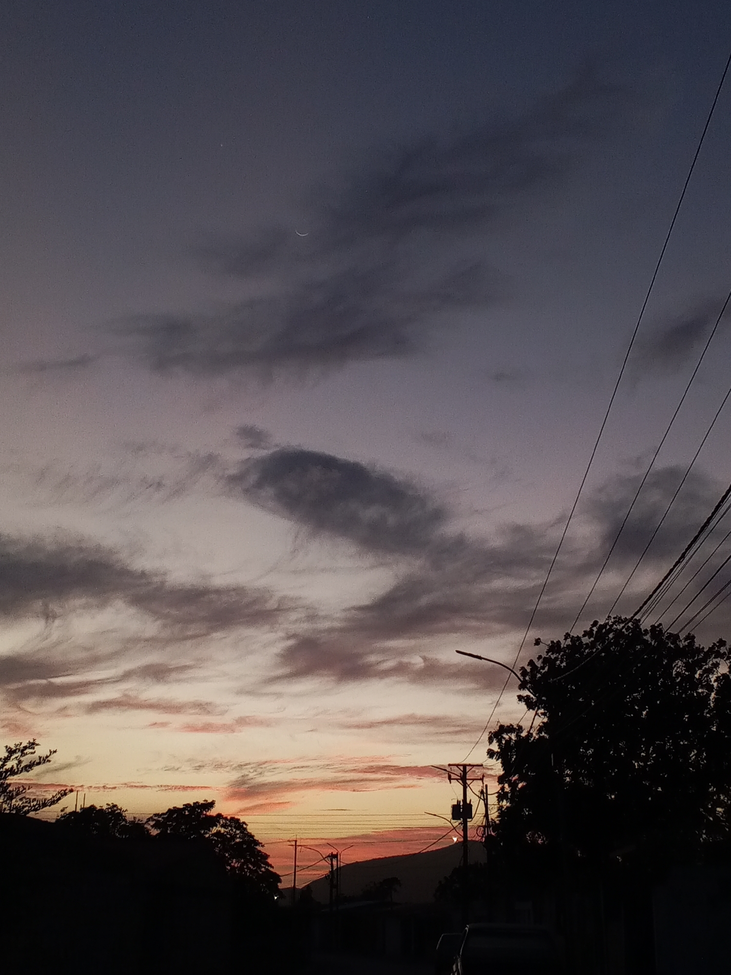 The image depicts a twilight scene in Altagracia, Venezuela. The sky transitions from a deep blue-gray to orange-pink hues near the horizon. Streaks of clouds are visible throughout the sky. A thin crescent moon and a small star are visible in the upper portion of the image. Electrical wires stretch across the right side of the frame. Silhouetted trees and a few power poles line the lower portion of the image. A streetlamp is visible to the right and a vehicle is barely discernible. The environment suggests a residential area during twilight.
