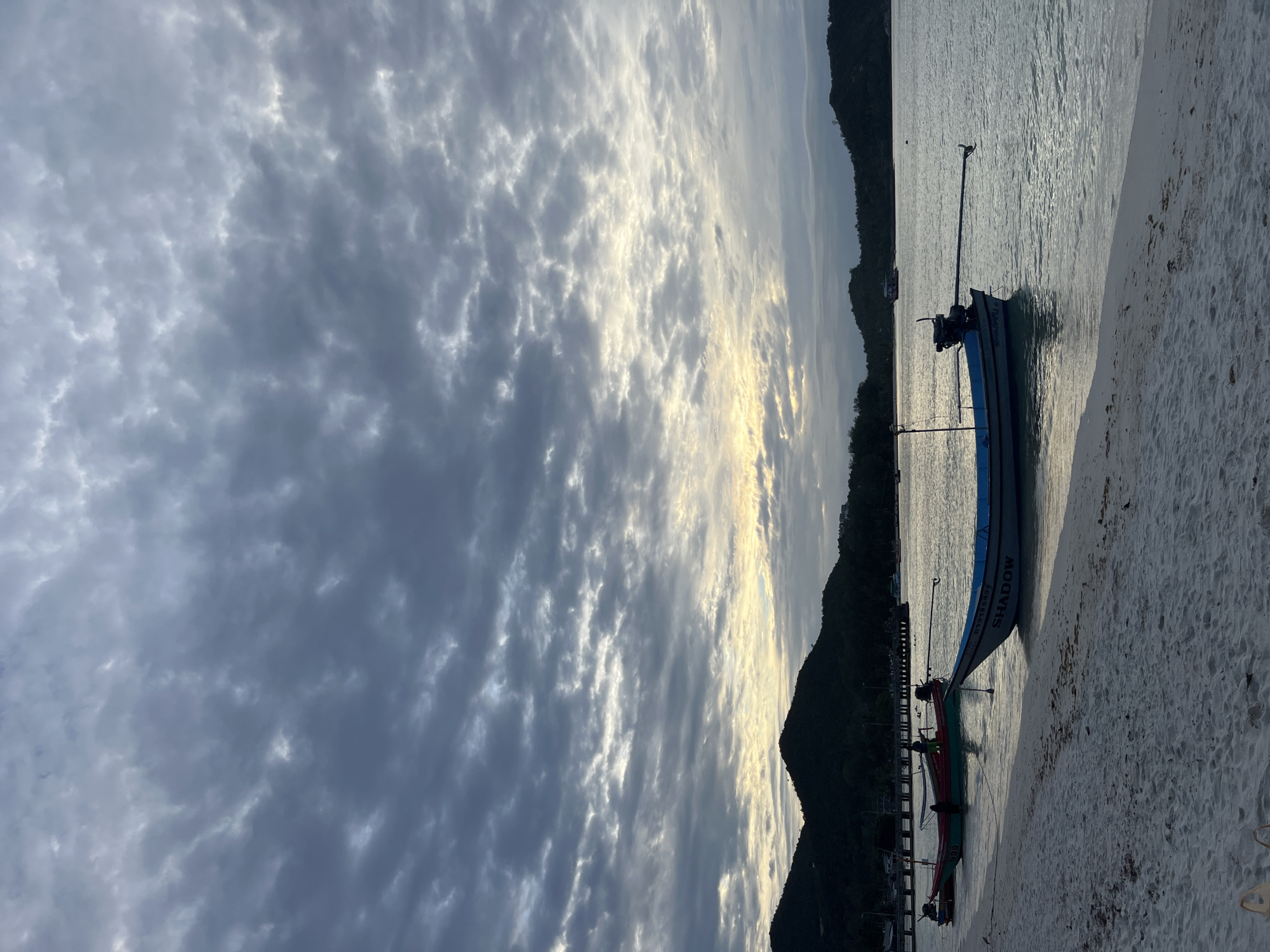 A tranquil beach scene unfolds under a cloudy sky, with gentle waves lapping at the white sand shore. Three longtail boats, commonly found in Southeast Asian waters, are moored near the beach. One boat, painted blue and white, prominently displays the name "SHADOW" on its side. Its engine, a visible detail, is poised at the stern, ready for use. Further out in the calm, shimmering water, other boats and structures dot the horizon, suggesting a populated coastal area. Dark, lush hills form a backdrop, their silhouettes softened by the diffused light of a sky filled with dramatic, textured clouds. The overall impression is one of peaceful serenity at the edge of the sea.