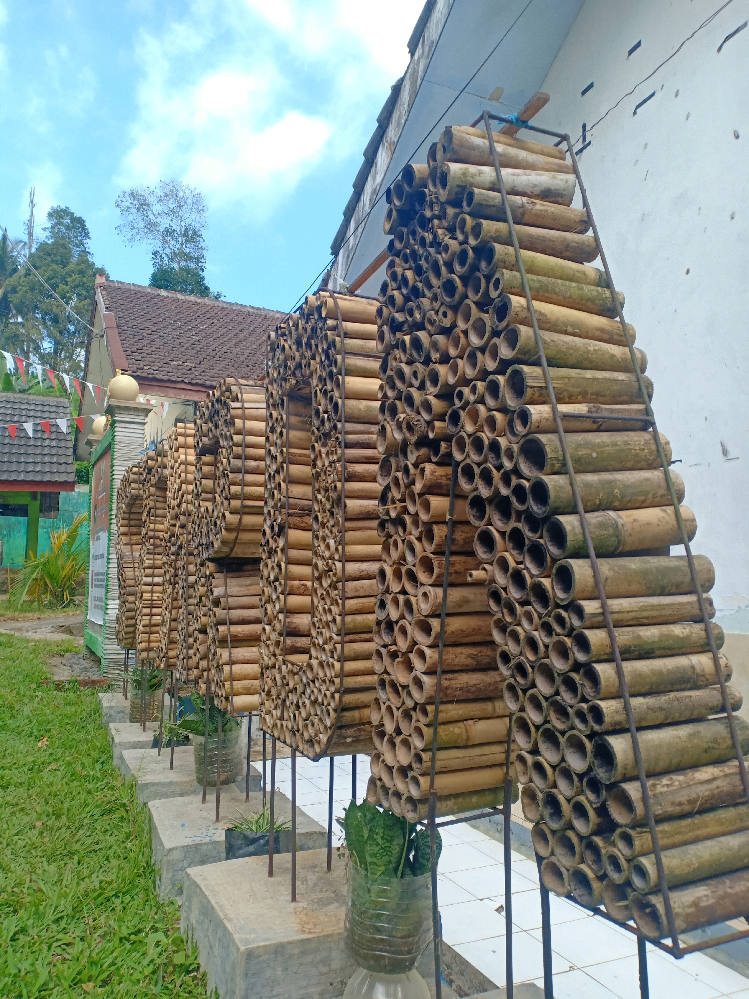 The image shows a decorative display made of stacked bamboo poles, forming the shapes of letters that spell "BHAKTI", positioned outdoors in front of a building with white walls and a tiled roof. The bamboo structure is supported by metal frames. In front of the bamboo display, there are several concrete blocks on the ground, each holding a plant in a plastic bottle. The area is surrounded by green grass and trees. There are also small triangular flags hanging on a fence to the left of the bamboo structure, and a sign on a post near the flags. The sky above is blue with scattered white clouds. This scene is located in Kaliuling, Indonesia. There are no people visible in the image.