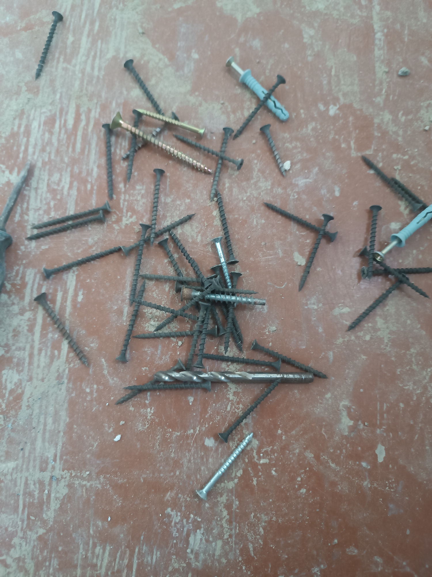 The image displays a collection of assorted screws and a drill bit scattered on a dusty, textured surface, possibly a wooden floor or workbench. The items are varied in size and material, including dark metal screws, a few gold-toned screws, and a light blue plastic anchor with a metal screw. The lighting suggests an indoor setting, possibly in a workshop or unfinished room. There are no people visible in the image, nor any indication of the specific location within Grinăuți, Moldova. The scene is static and appears to be a simple arrangement of hardware.