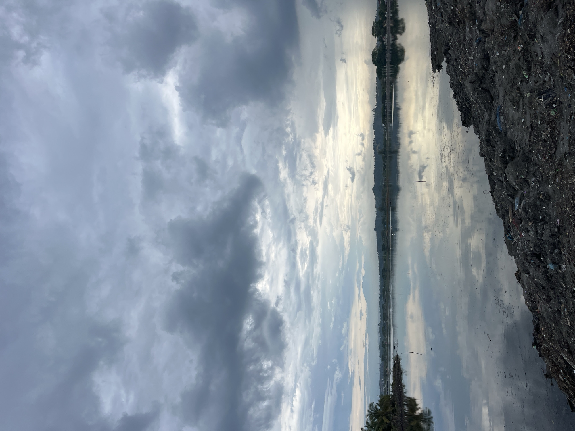 The image depicts a body of water near Lhokseumawe, Indonesia, under a cloudy sky. The sky has varying cloud densities, ranging from light to dark grey. The water reflects the sky, creating a mirror effect. On the right side of the image, there is a substantial pile of dark-colored material, possibly waste or debris. Vegetation, including palm trees, is visible in the foreground. In the distance, across the water, there is a strip of land with structures or buildings.