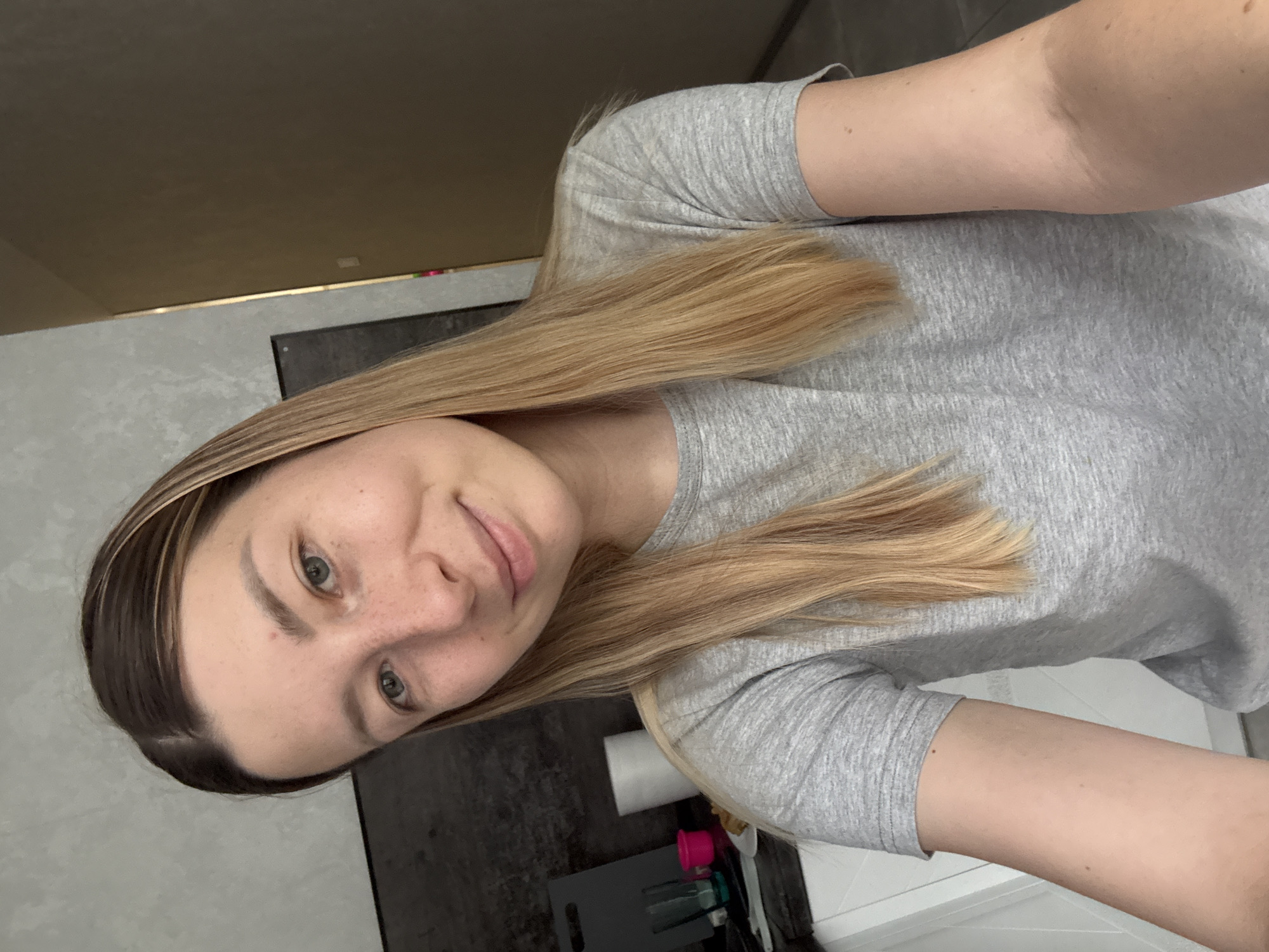 A young woman with blonde hair and green eyes, wearing a grey t-shirt, is taking a selfie in what appears to be a domestic setting. She has freckles across her nose and cheeks and is smiling. In the background, on a counter, are various items including what looks like a white paper towel dispenser, a small pink object, and some green cups. The wall behind her has a textured grey finish. The overall impression is a casual, indoor environment. There is no indication of her being in Moscow, Russia.
