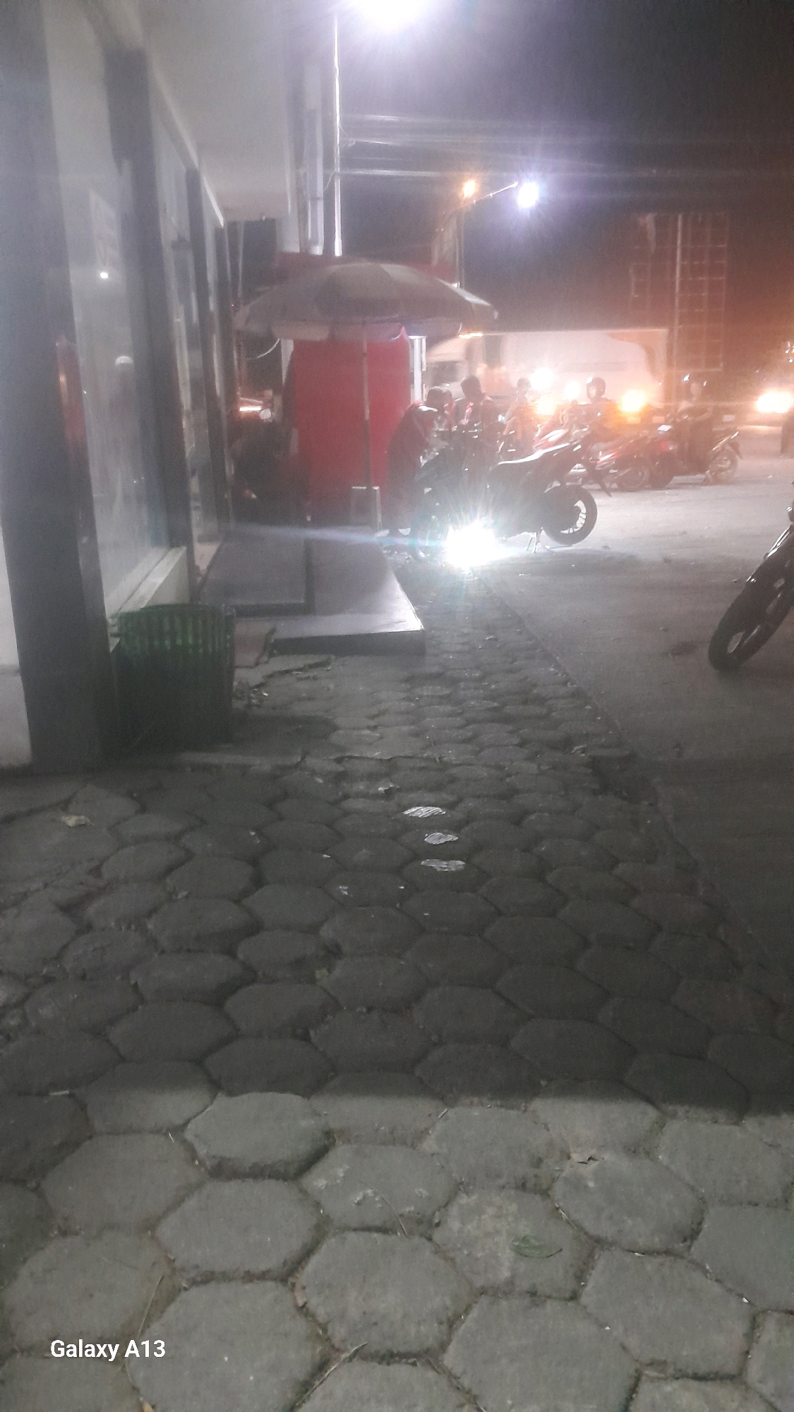 The image depicts a nighttime scene on a street in Jekulo, Indonesia, with a focus on a sidewalk paved with hexagonal stones. In the background, several motorcycles are parked, with one in the foreground emitting a bright headlight glare. A red cylindrical object, possibly a large container or pillar, is visible to the left, topped with an umbrella. The surroundings suggest an urban environment, with lights from buildings and street lamps illuminating the area. A green trash bin is also present near the building on the left. The overall atmosphere is dimly lit, characteristic of a street scene at night.