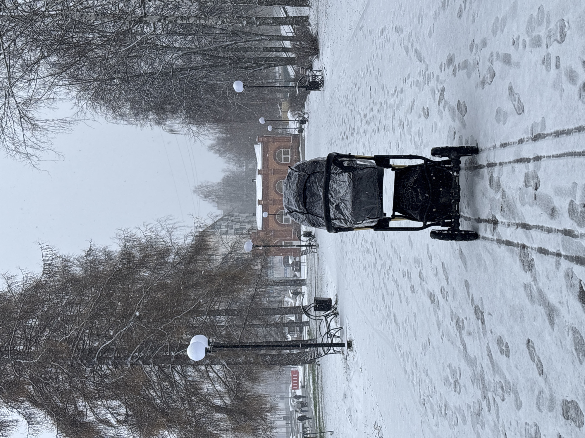 A stroller sits on a snow-covered path in Chaikovsky, Russia. The path is flanked by bare trees and lampposts, leading to a brick building in the distance. Footprints and stroller tracks mark the snowy ground. Snow falls throughout the scene. Benches are visible along the path.