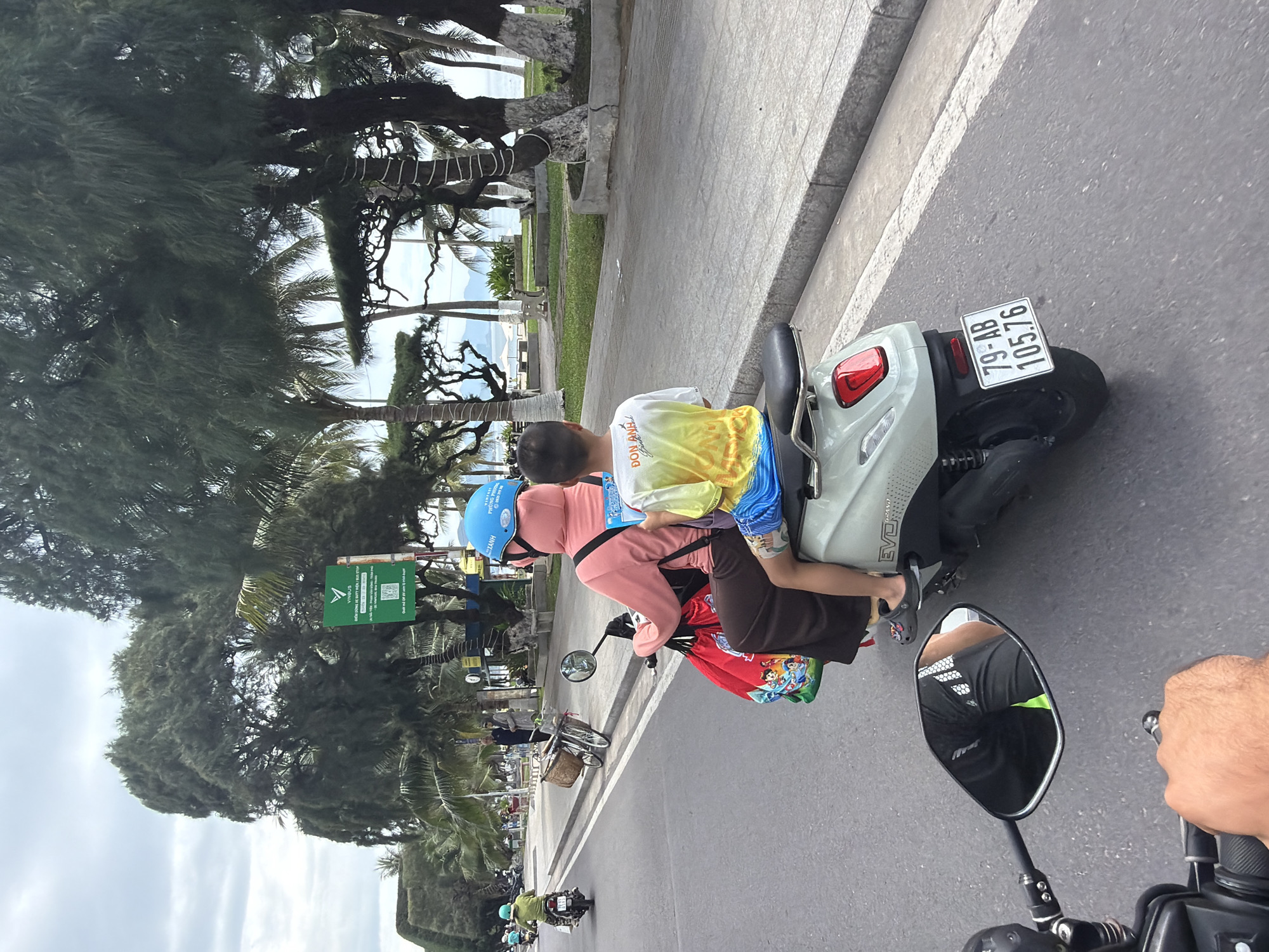 The image captures a street scene in Nha Trang, Vietnam, with a focus on a scooter carrying two individuals. The driver, wearing a pink hooded garment and a blue helmet, is operating the scooter. A child, dressed in a yellow and green t-shirt and blue shorts, sits behind the driver. The scooter has a license plate reading "79-AB 105.76". A red bag with cartoon characters is attached to the back of the scooter. The scooter is traveling on a paved road alongside a sidewalk. In the background, there are trees, palm trees, and a glimpse of the ocean. Signs are visible on the side of the road, including a green sign with Vietnamese text and a QR code. Other vehicles and pedestrians are present in the distance, indicating a typical urban environment. The overall atmosphere suggests a daytime scene in a coastal city.
